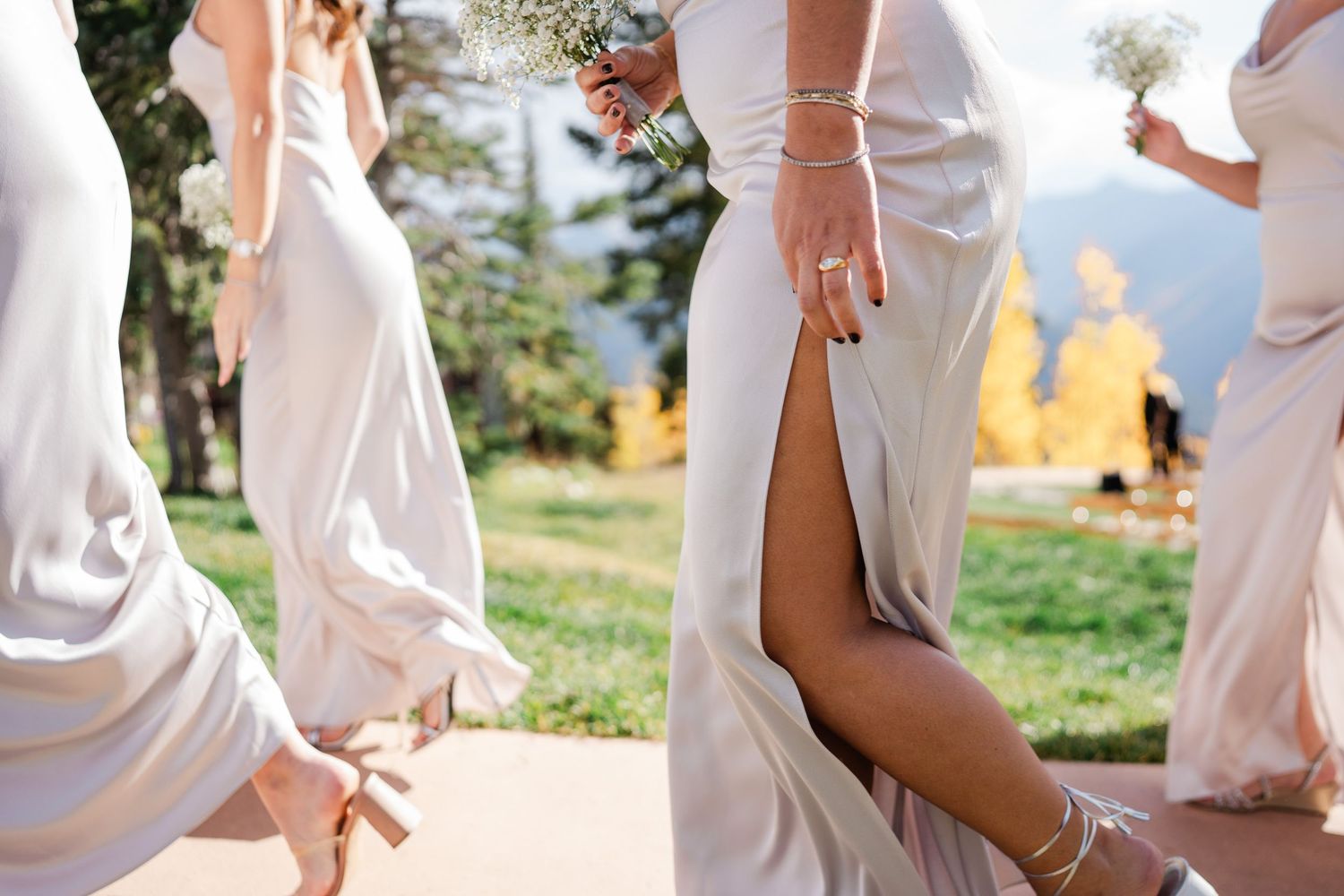 Close-up of bridesmaids in champagne slit gowns holding baby's breath bouquets walking outdoors at a mountain wedding.