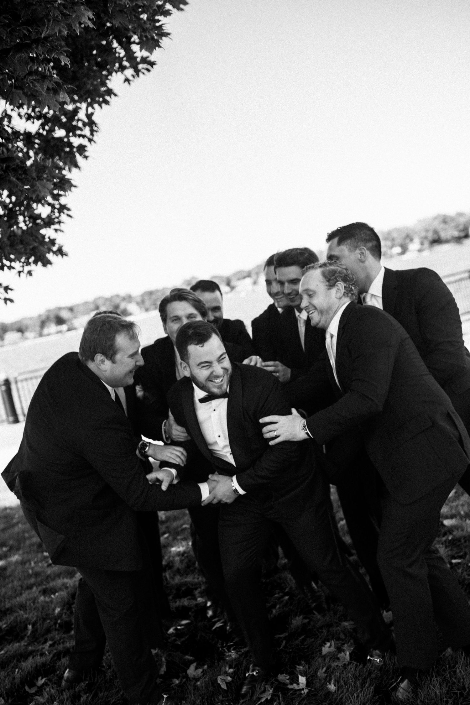 Black and white photo of groom laughing with groomsmen in tuxedos outdoors near a waterfront.