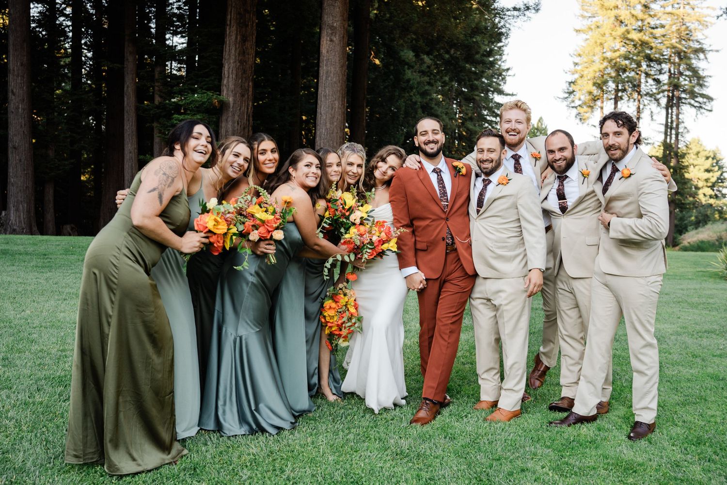 Wedding party posing outdoors in forest setting, bridesmaids in sage green gowns, groomsmen in tan suits, bride and groom centered.