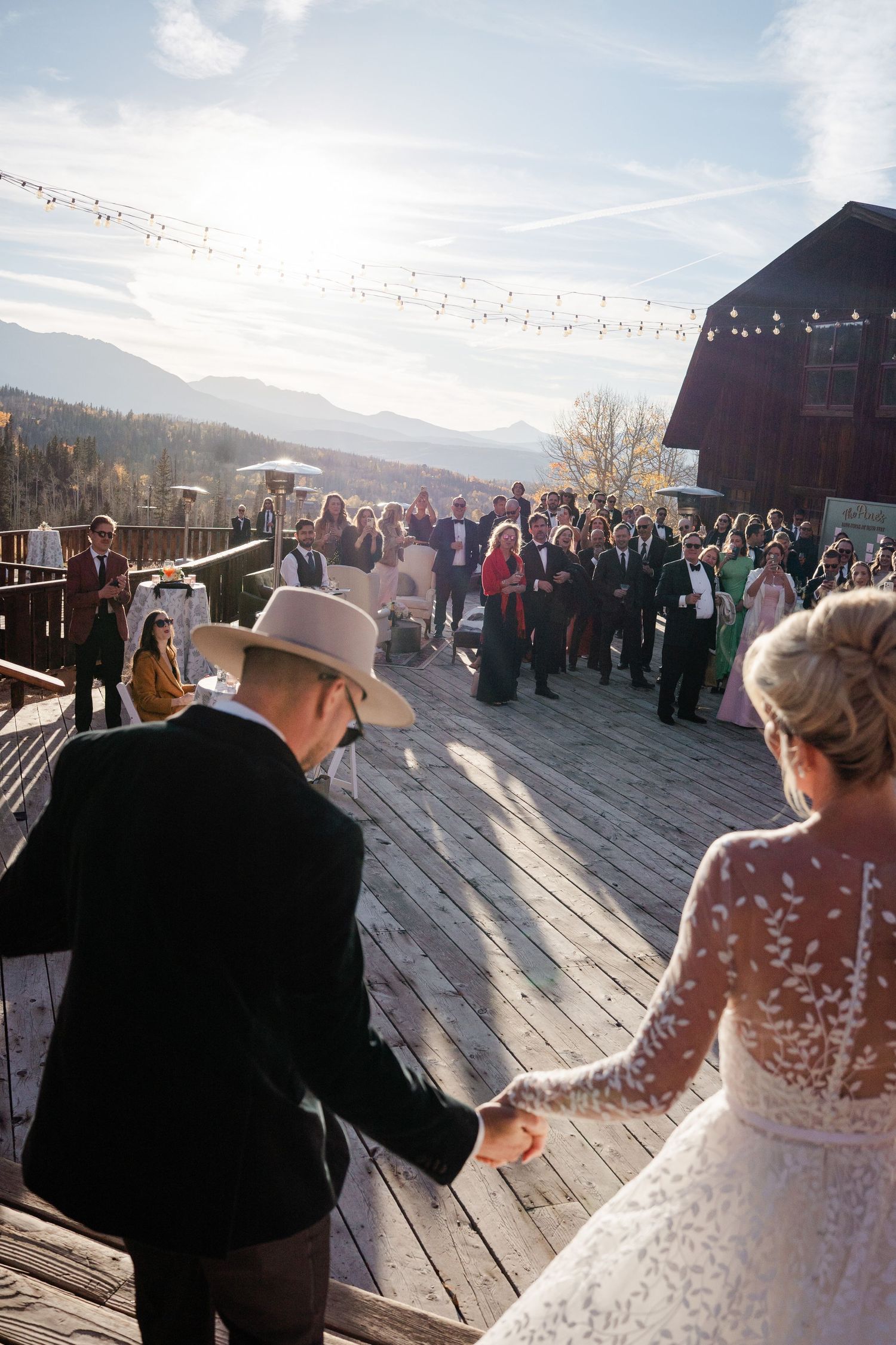 Bride and groom entering outdoor wedding reception on a wooden deck with mountain scenery.