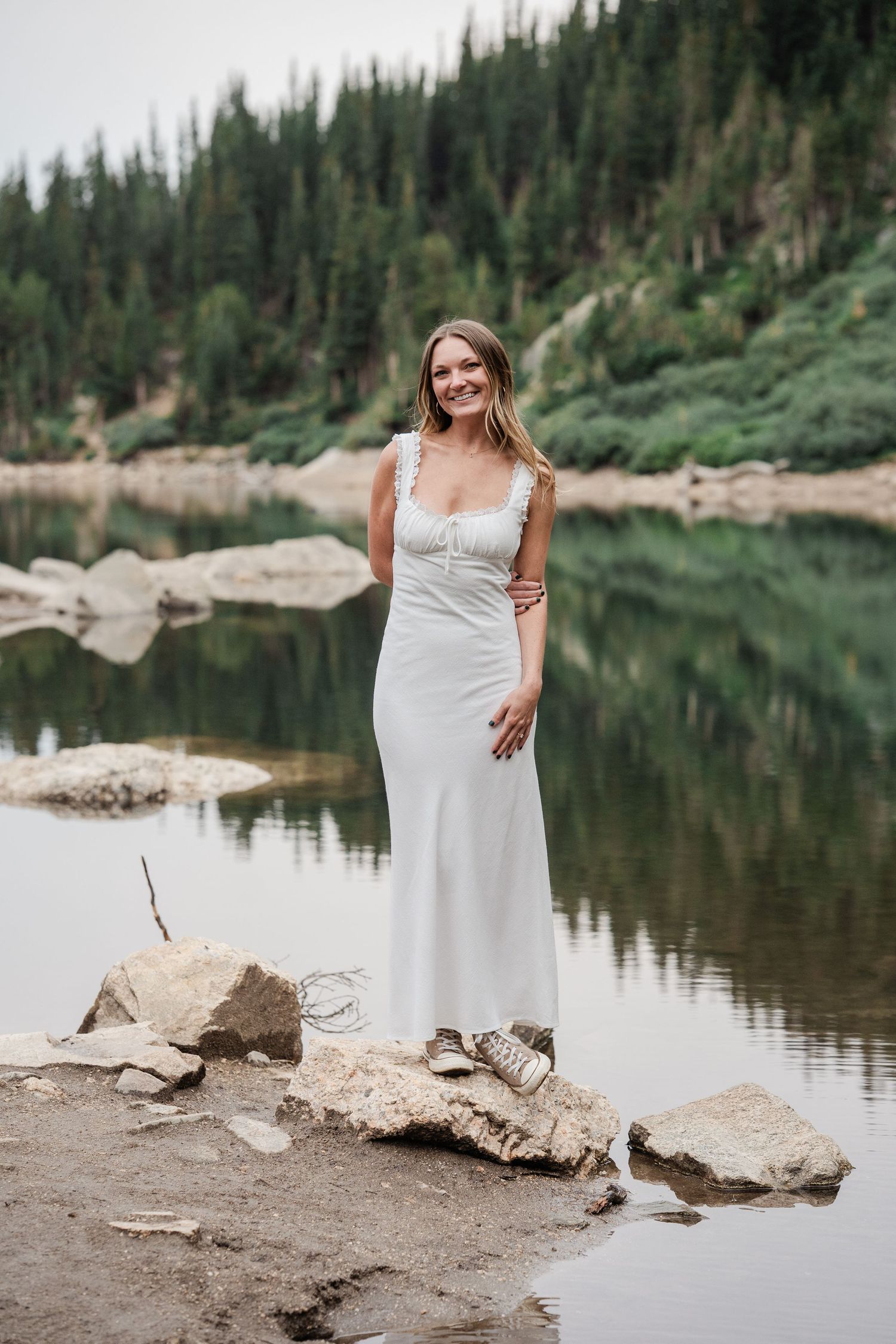 Smiling bride in white lace dress standing on rocks by a mountain lake surrounded by pine trees.