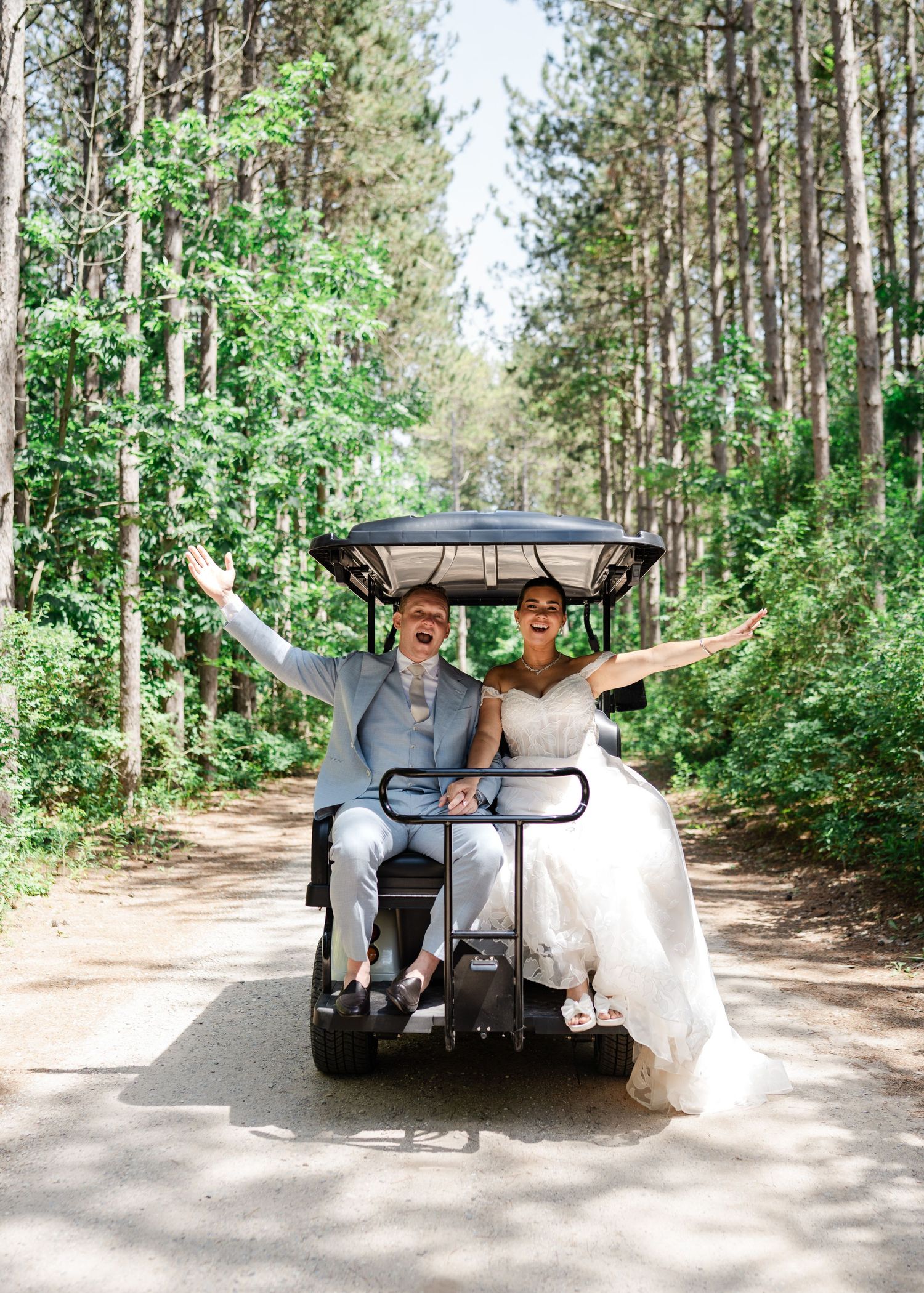 Laughing newlyweds ride a golf cart through a pine forest road with arms raised in celebration.