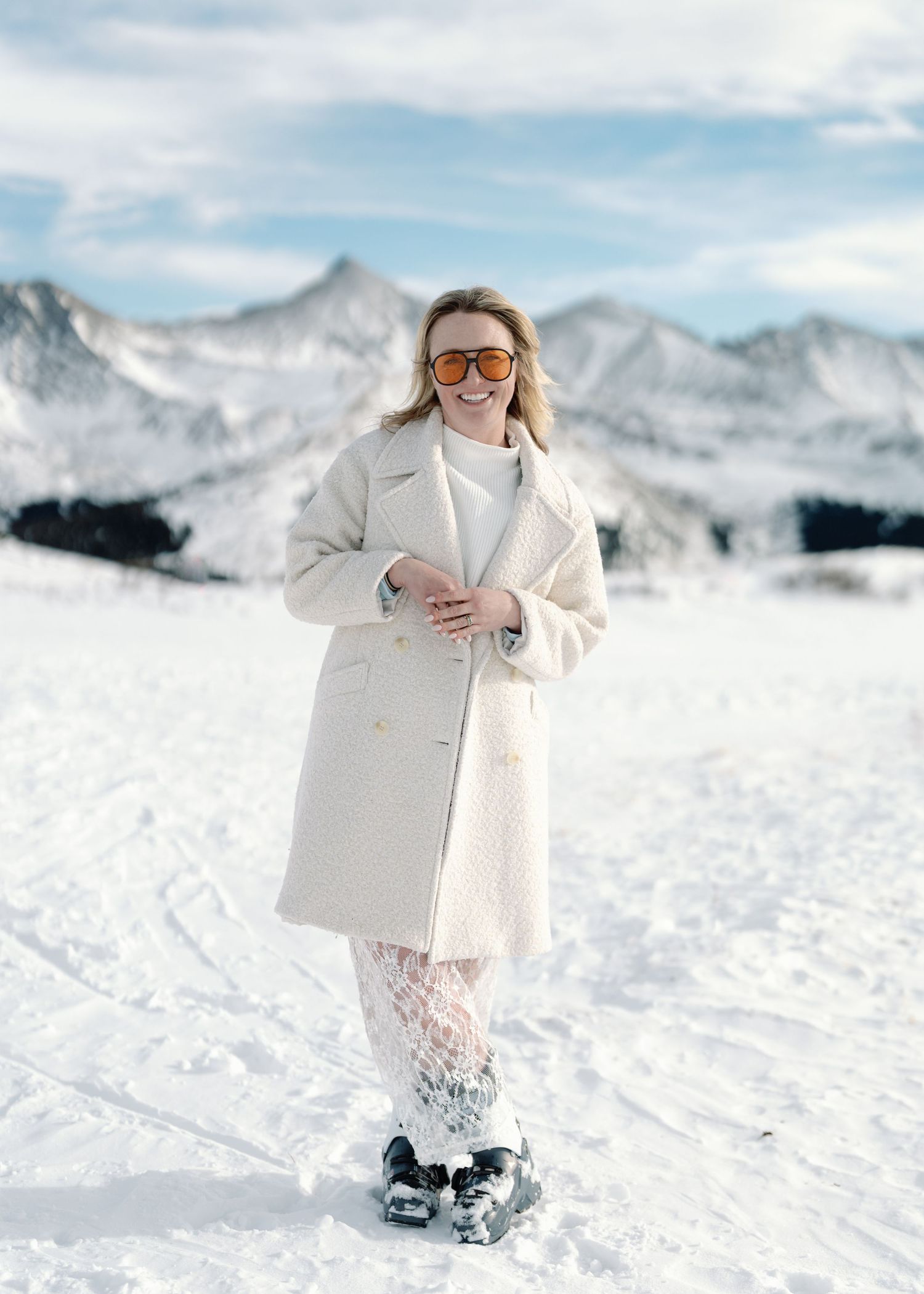 Smiling blonde woman in white coat and orange sunglasses stands in deep snow with mountain views.