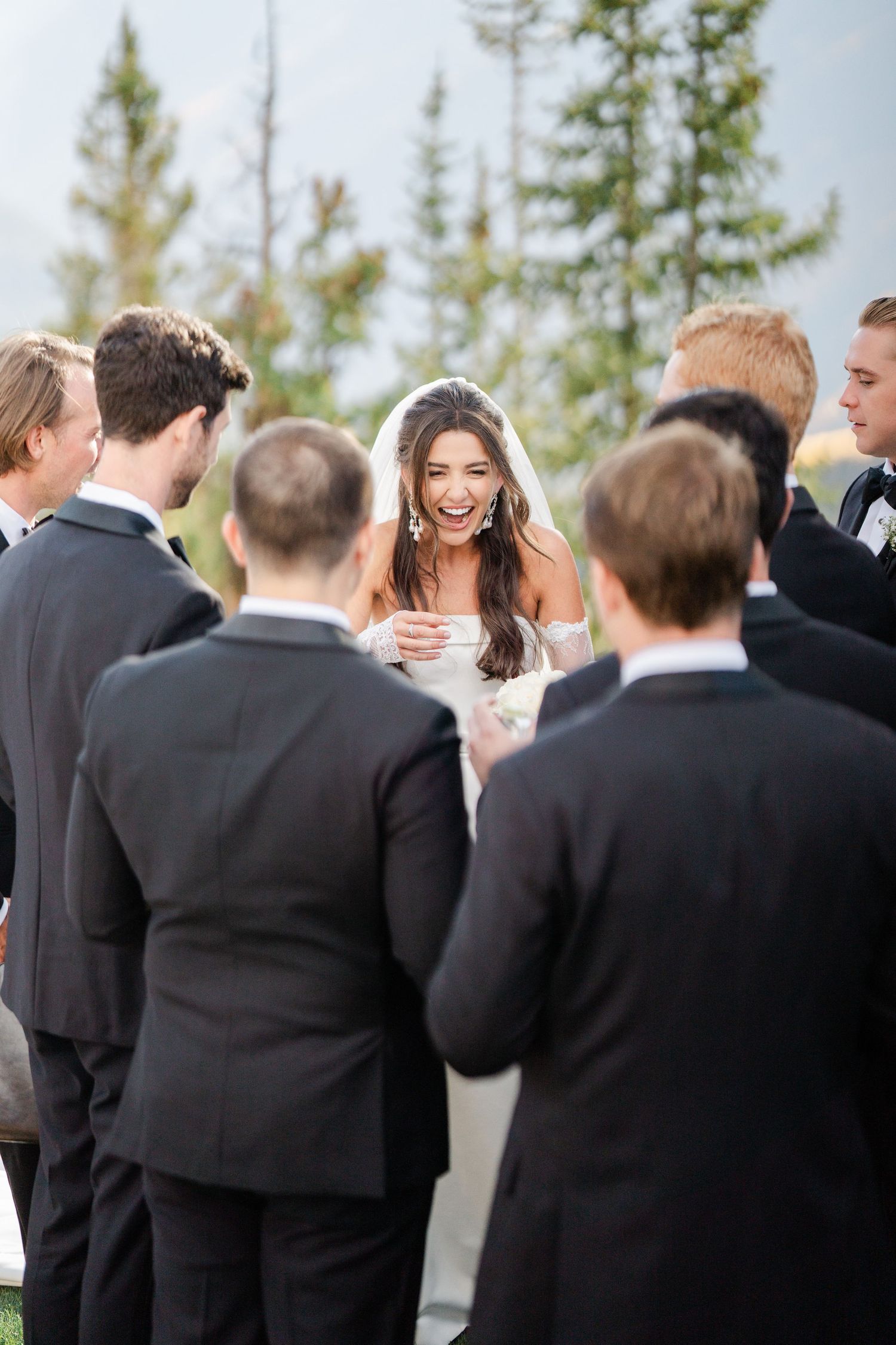 Joyful bride in white veil laughs surrounded by groomsmen in black suits at an outdoor mountain wedding.