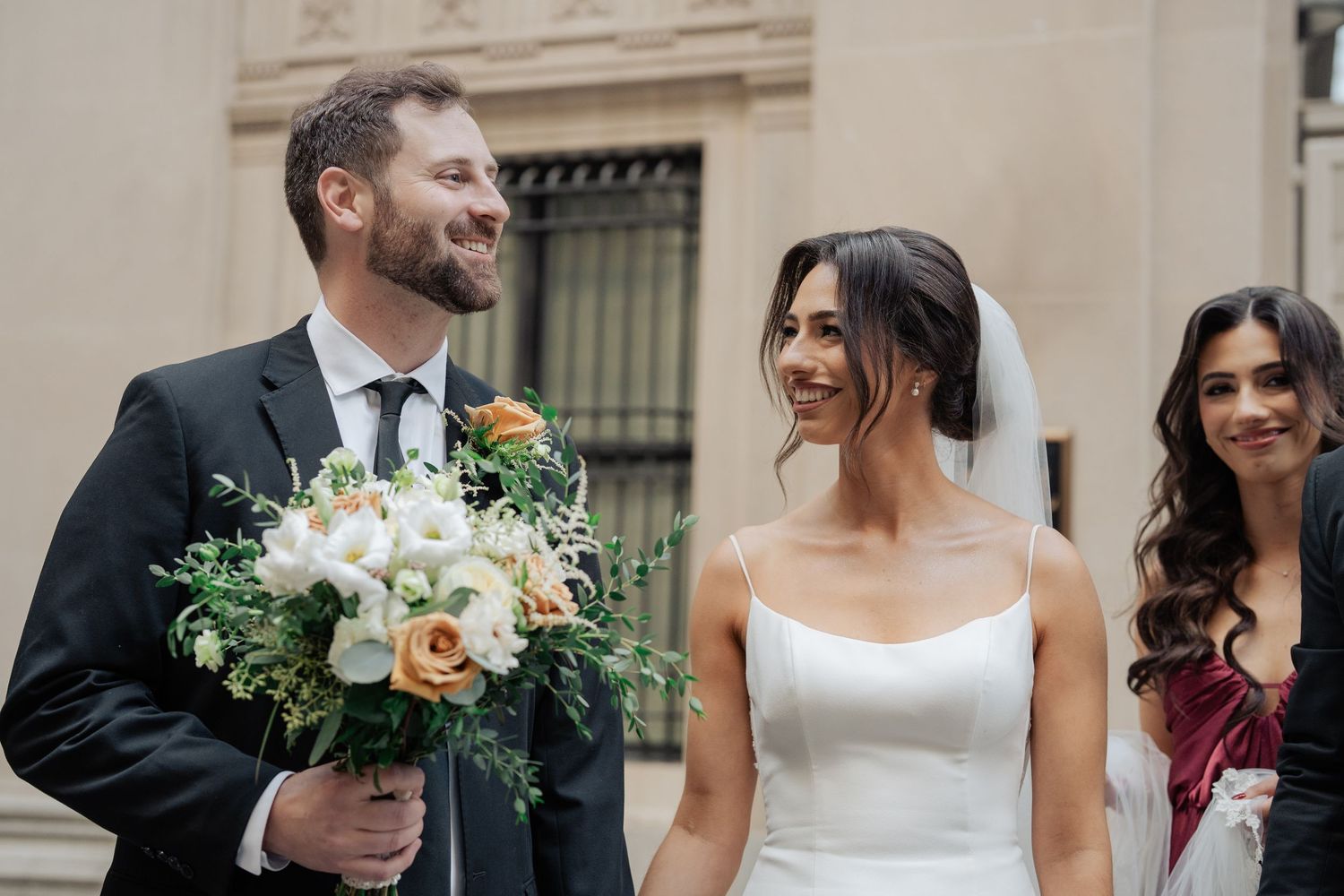 Smiling bride in white gown holding peach and white bouquet with groom in black suit at outdoor ceremony.