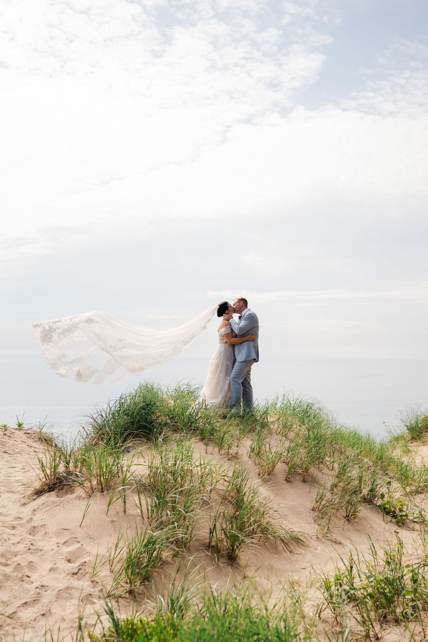 Bride and groom kissing on sand dune with veil blowing in wind.