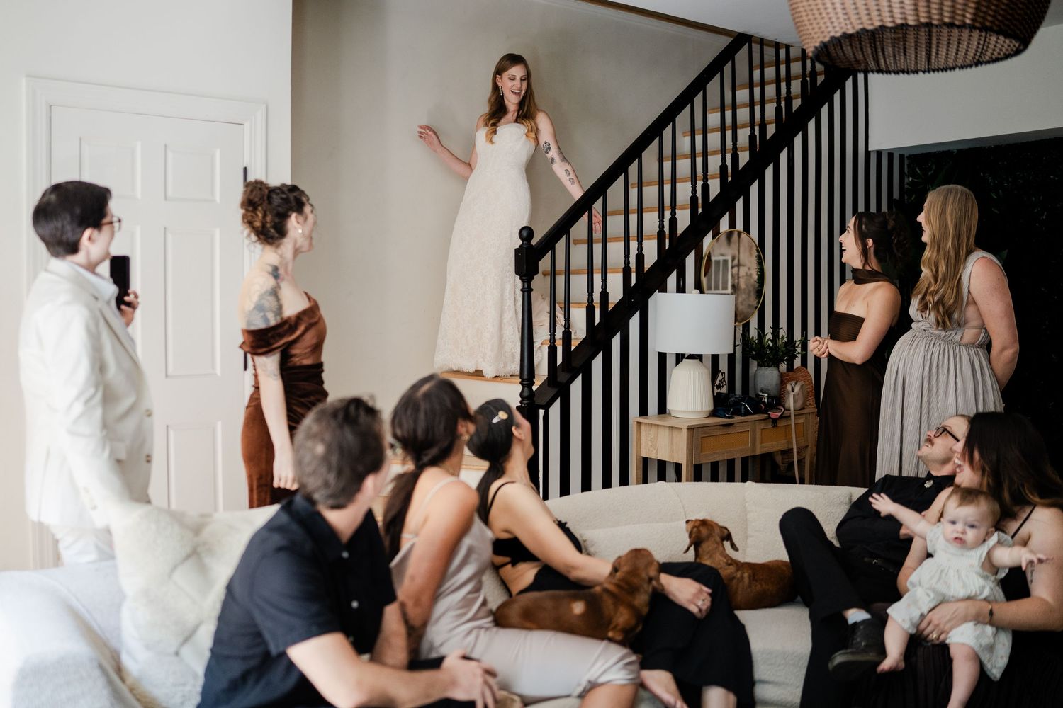 Bride in white gown stands on staircase revealing look to family and friends gathered below.