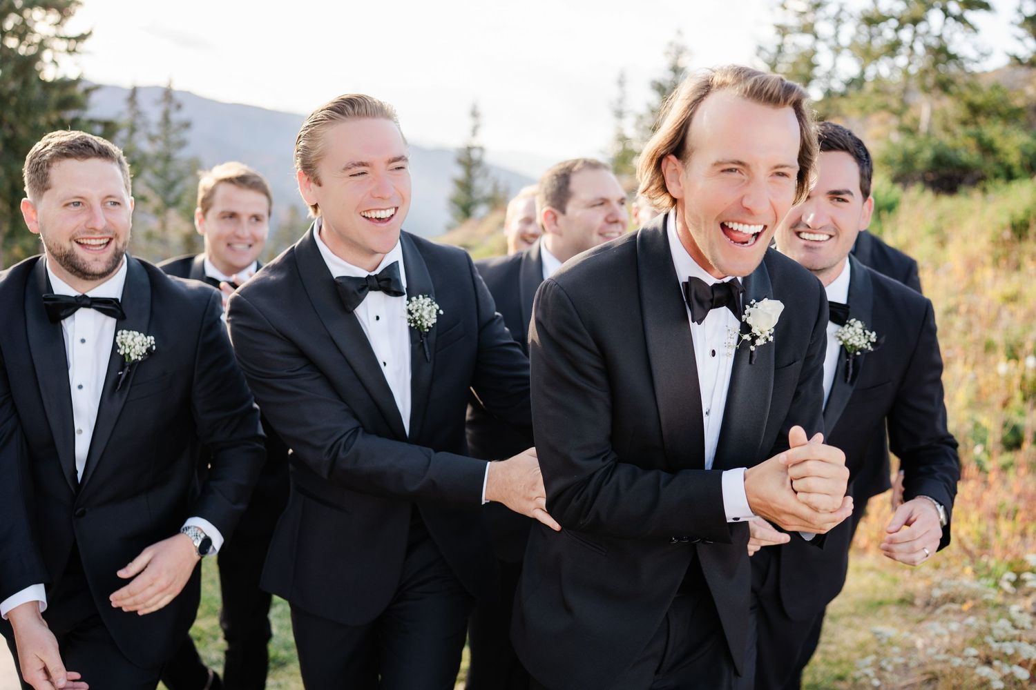 Groom and groomsmen in black tuxedos laughing together in a scenic outdoor mountain setting.