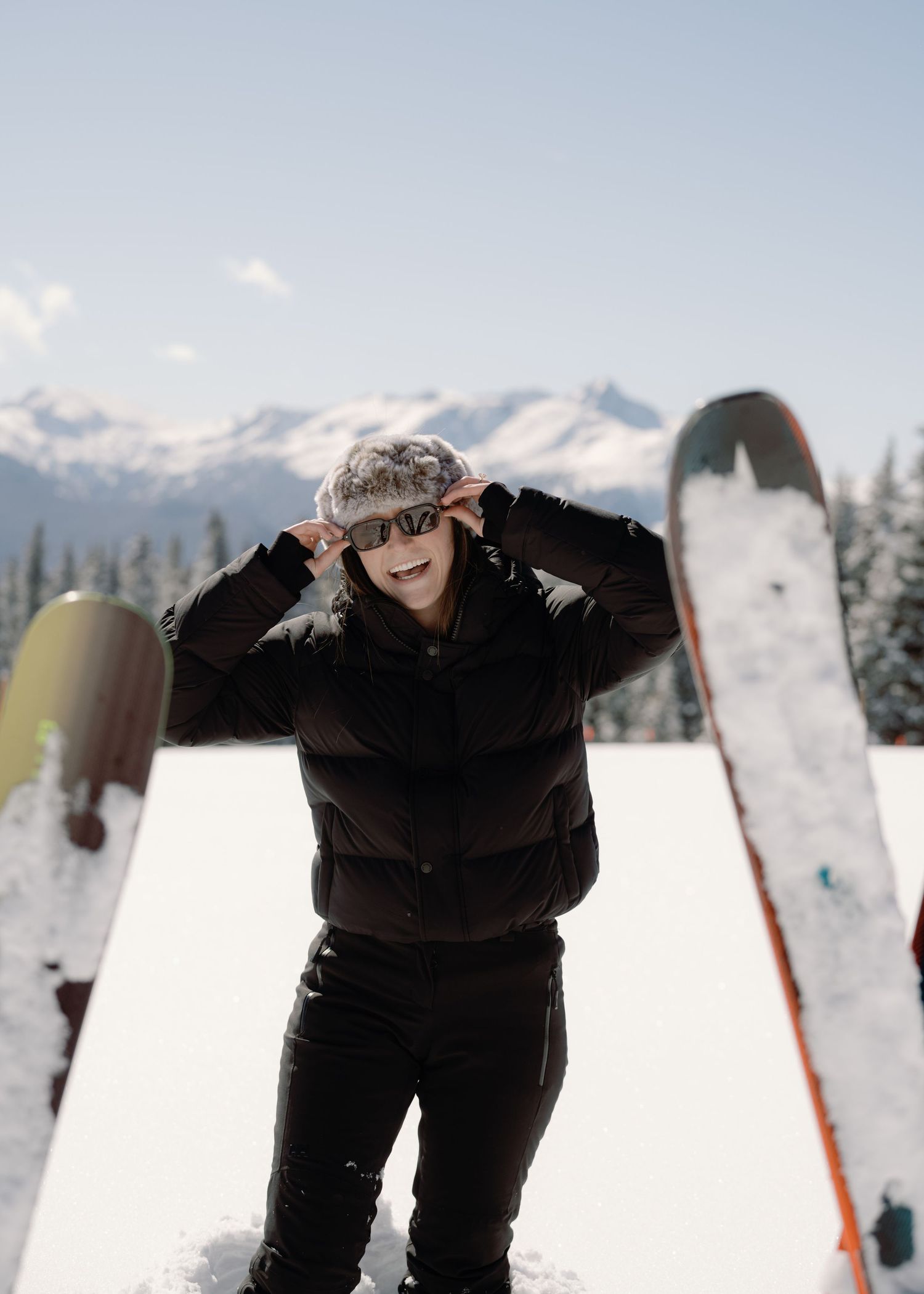 Woman in black puffer jacket and fur hat smiling while adjusting sunglasses on snowy mountain.