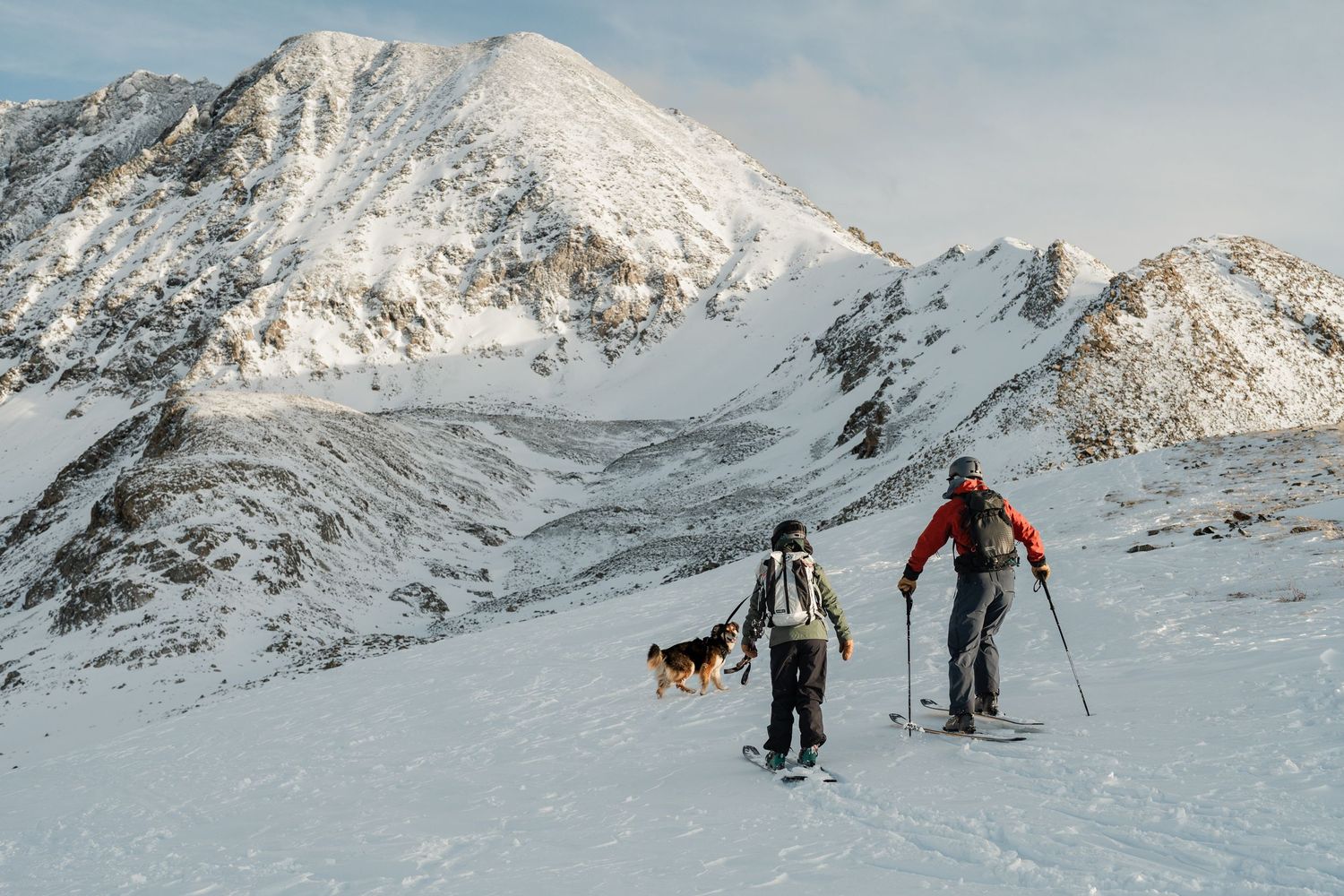 Two backcountry skiers and a dog ascending snowy mountain terrain with dramatic peaks in background.
