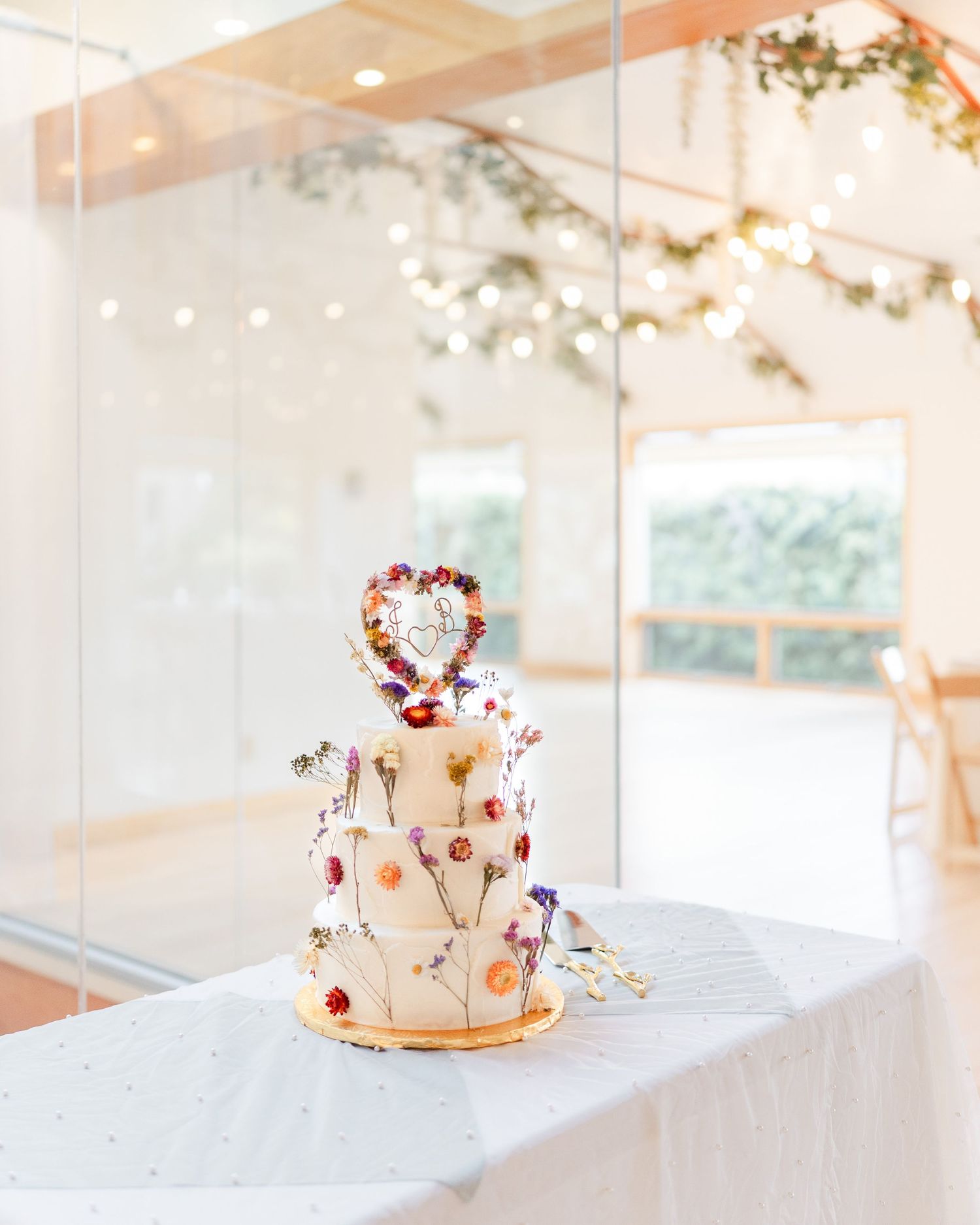 Elegant three-tier white wedding cake adorned with colorful pressed flowers and heart topper displayed on white table.