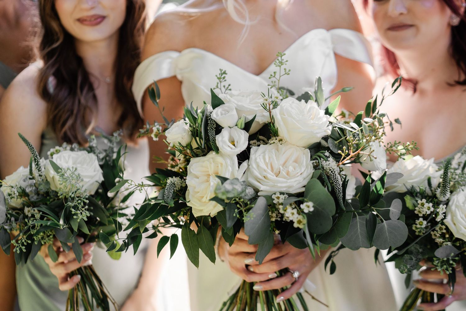 Bride and bridesmaids holding elegant white rose and greenery bouquets at a classic wedding ceremony.