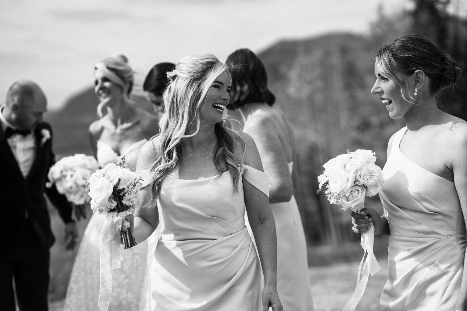Bride and bridesmaids in white dresses walking outdoors holding white bouquets in black and white photo.