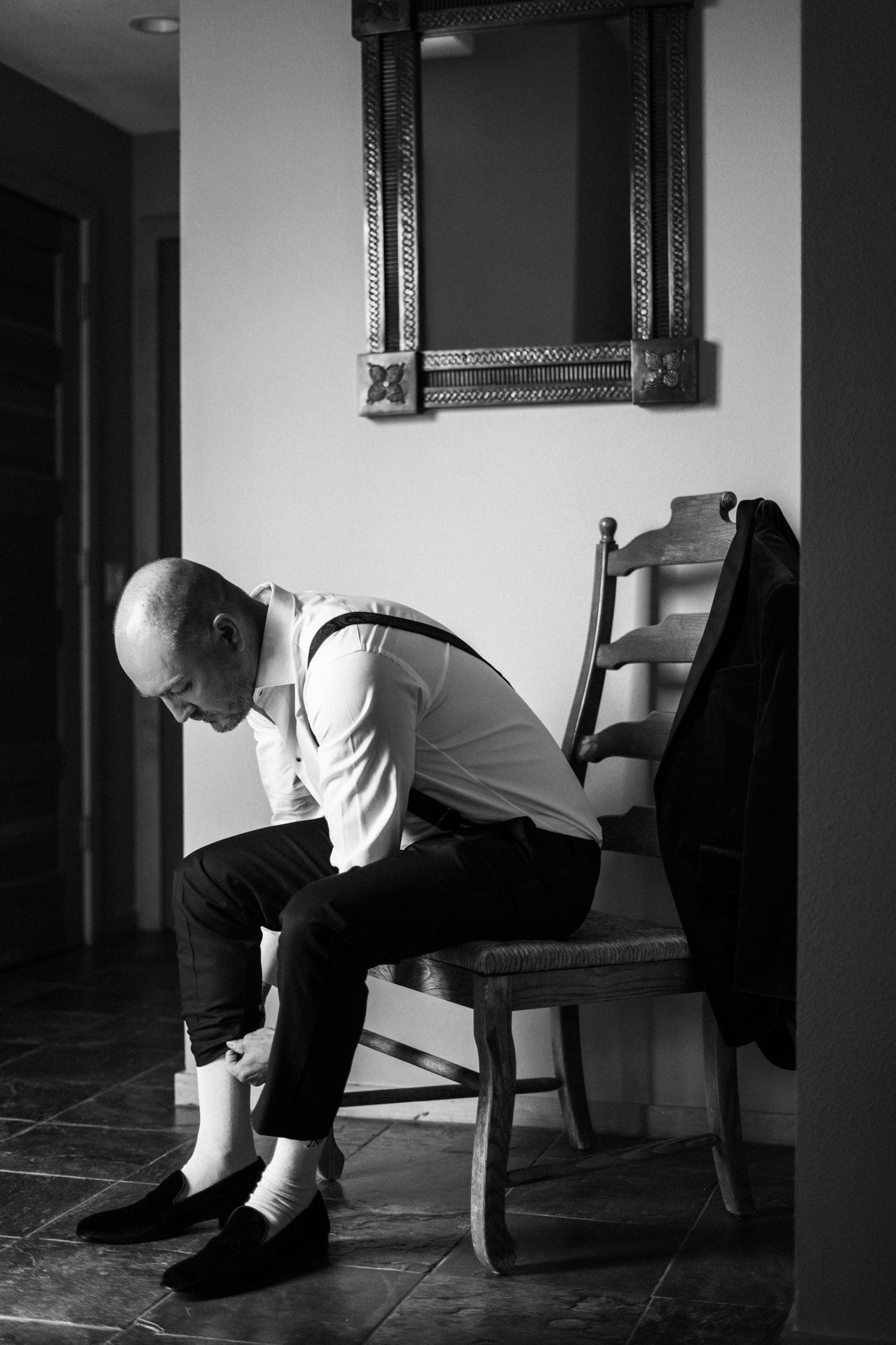 Black and white photo of a bald groom sitting on a chair getting dressed before his wedding.