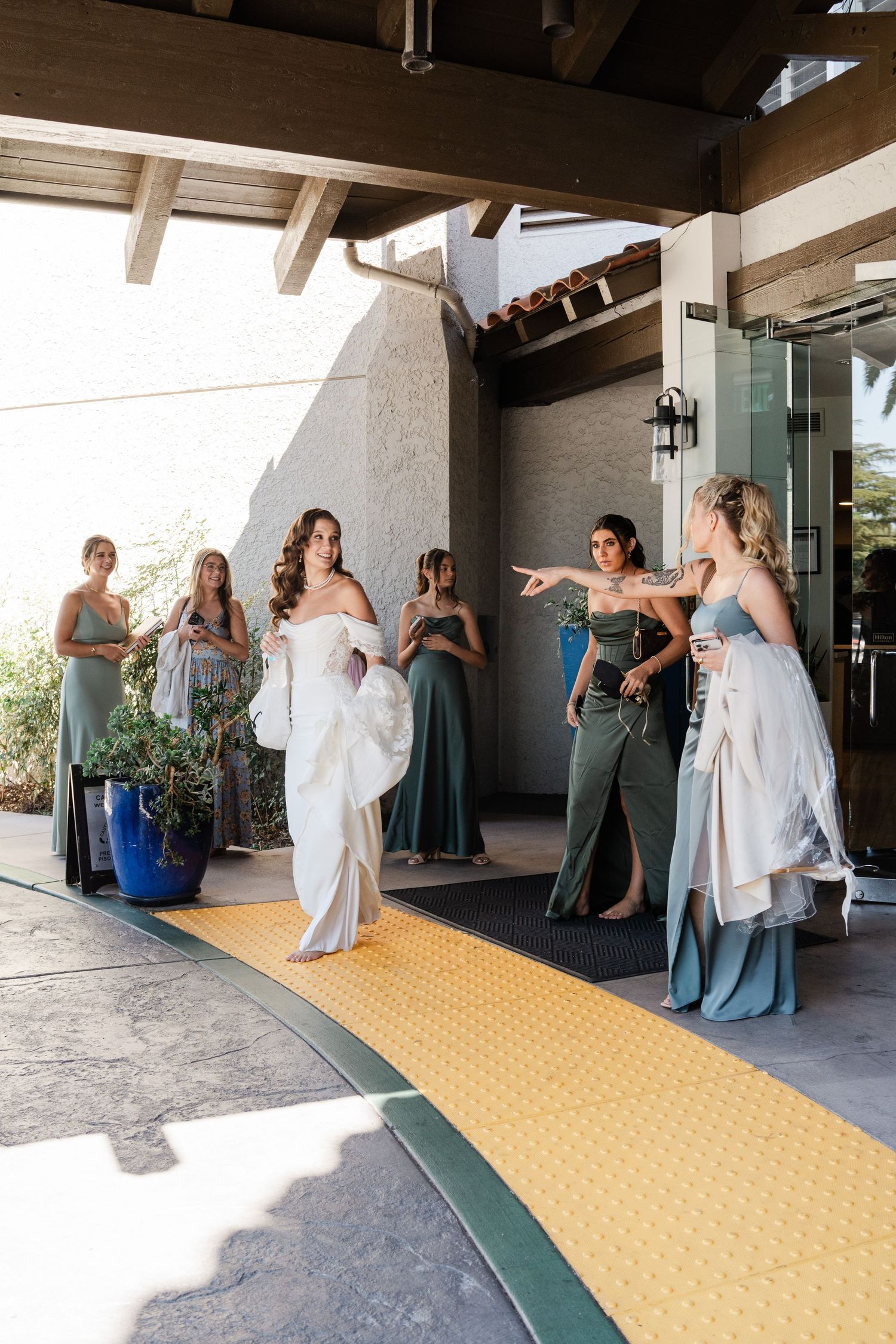 Bride in white gown greeted by bridesmaids in sage and green dresses at a resort entrance.