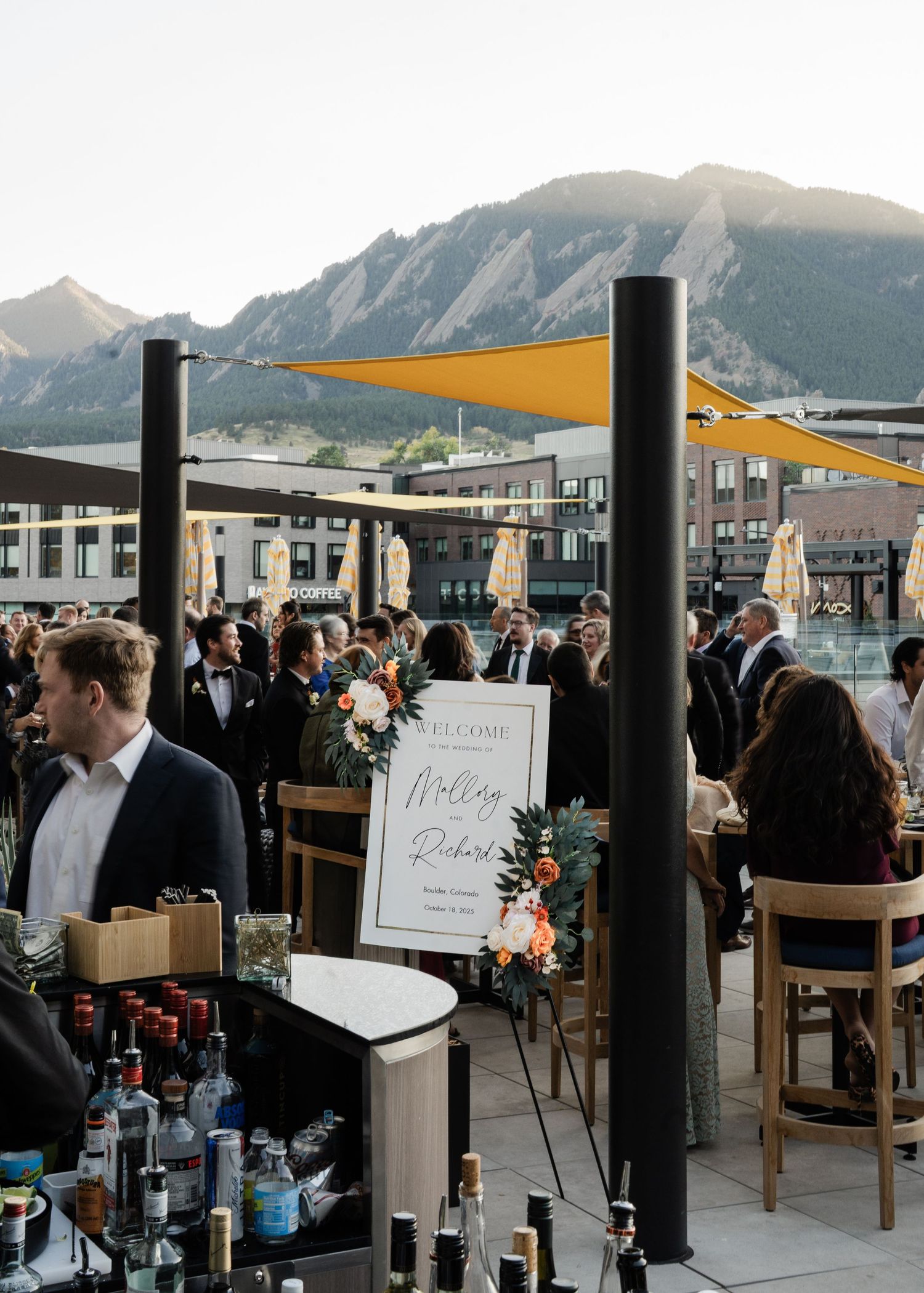 Outdoor rooftop wedding cocktail hour with mountain views, welcome sign, and elegantly dressed guests.