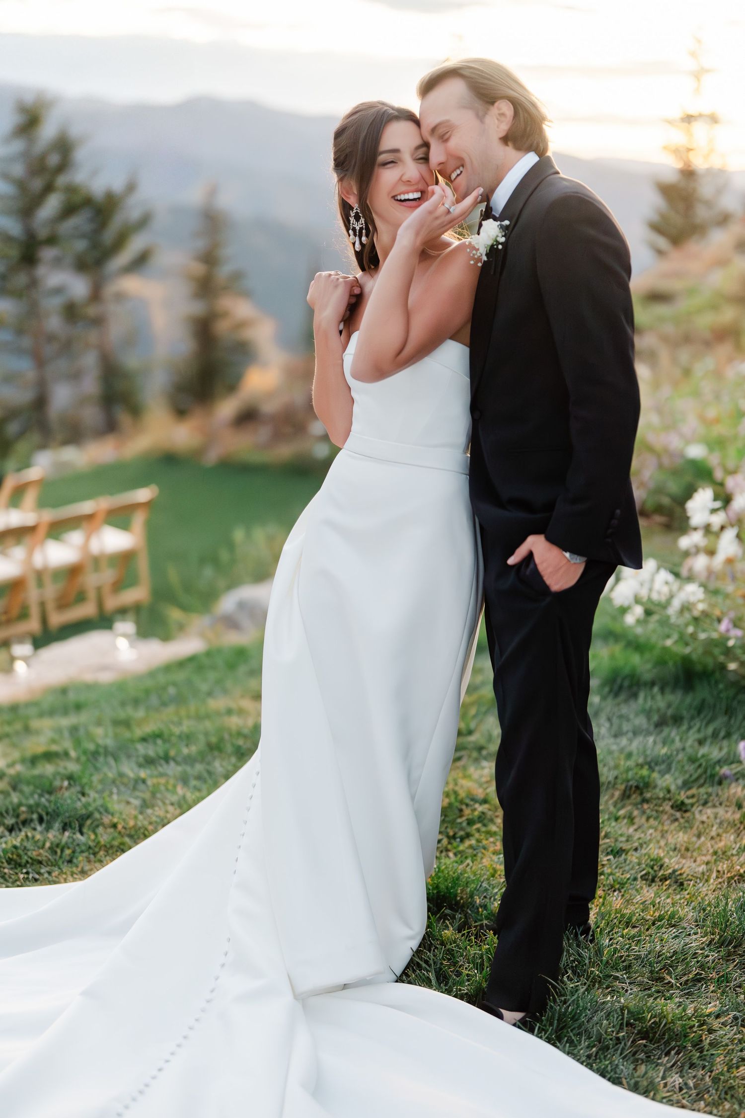 Smiling bride in white gown and groom in black tux pose on a mountain overlook at sunset.
