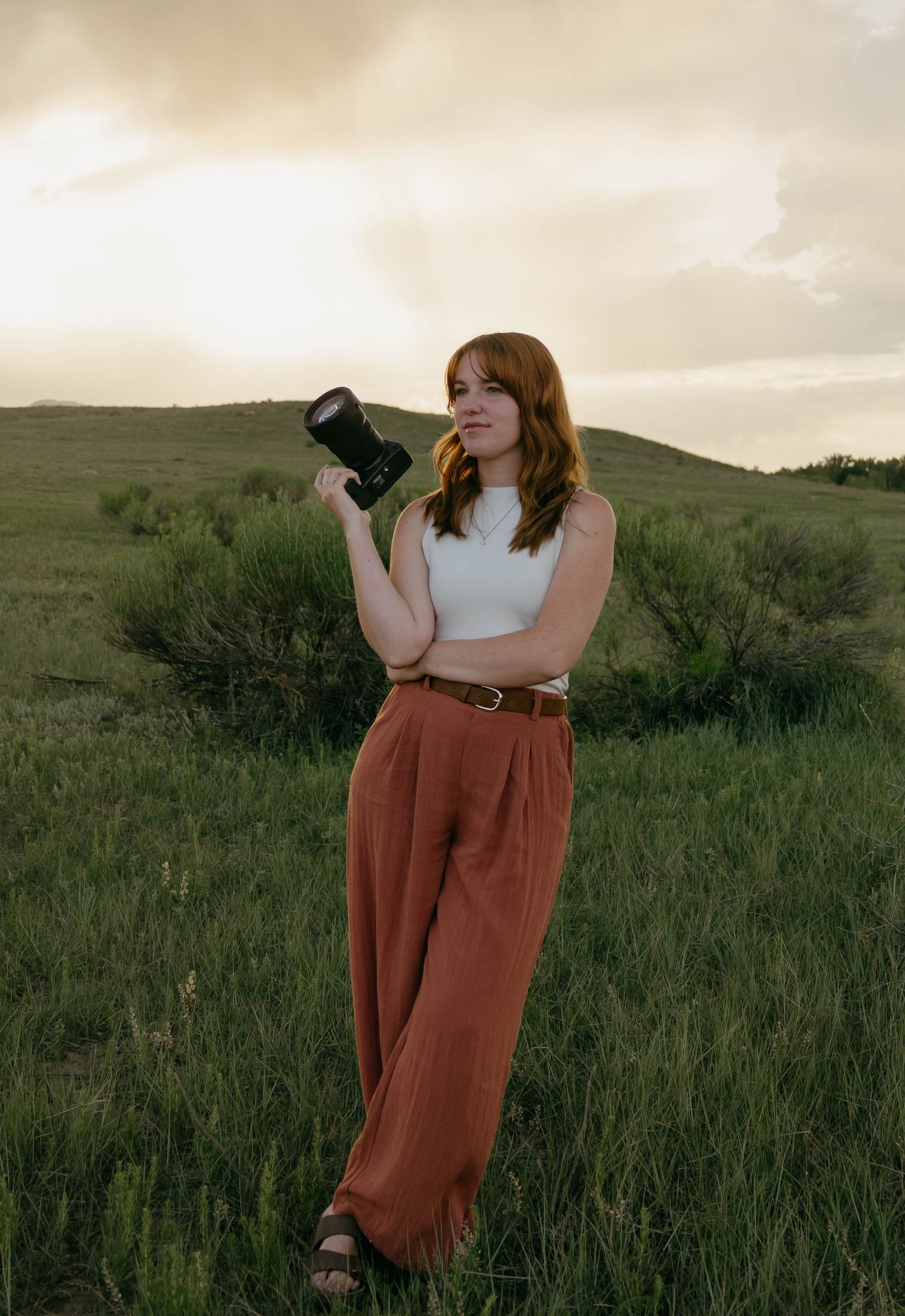 Red-haired woman holding a camera standing in a green field at sunset wearing rust-colored wide-leg pants.