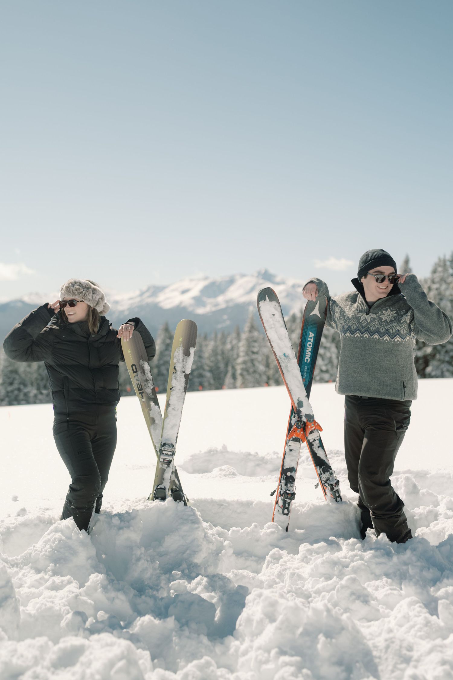 Couple posing with skis on snowy mountain wearing sunglasses and winter outfits on clear day.