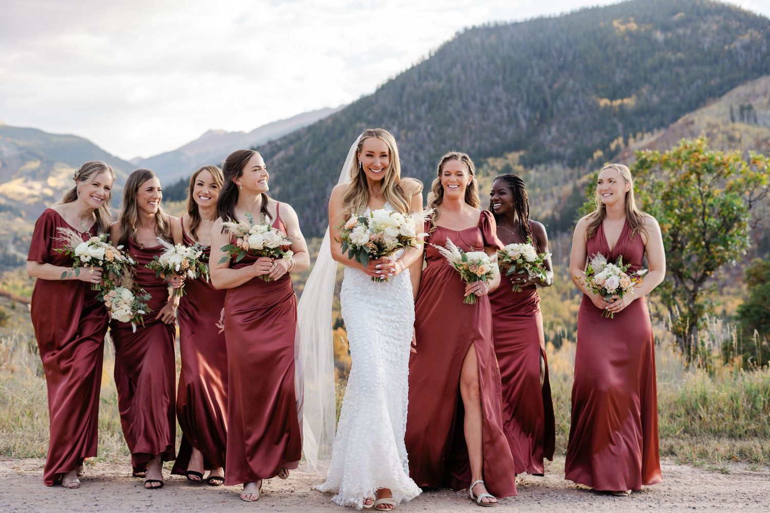 Bride in white gown walks with bridesmaids in burgundy satin dresses against a mountain landscape.