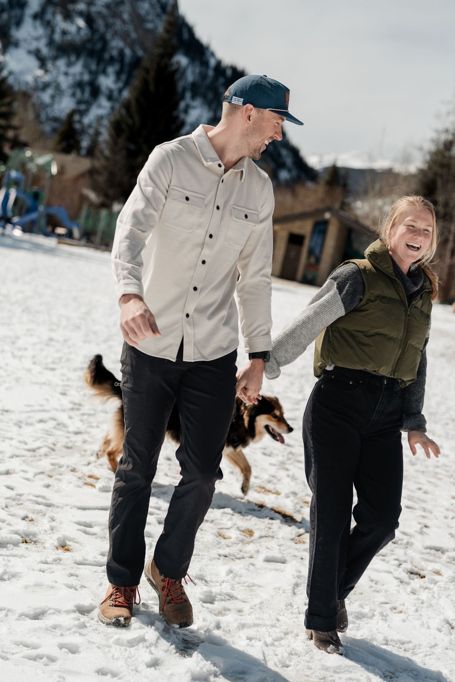 Laughing couple walking with dog in snowy mountain town on a sunny winter day.