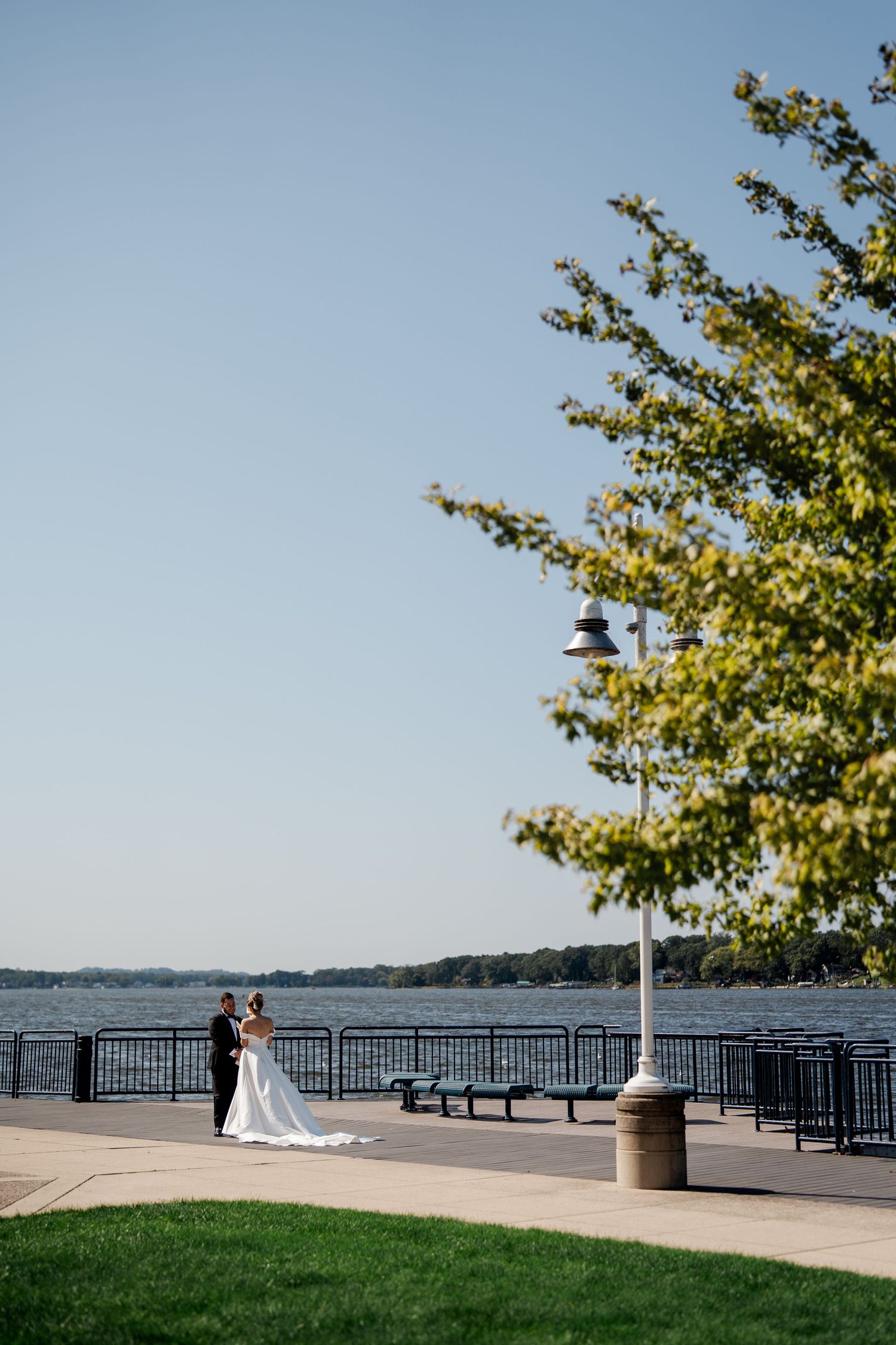 Bride and groom share a romantic embrace on a scenic waterfront promenade under a clear blue sky.