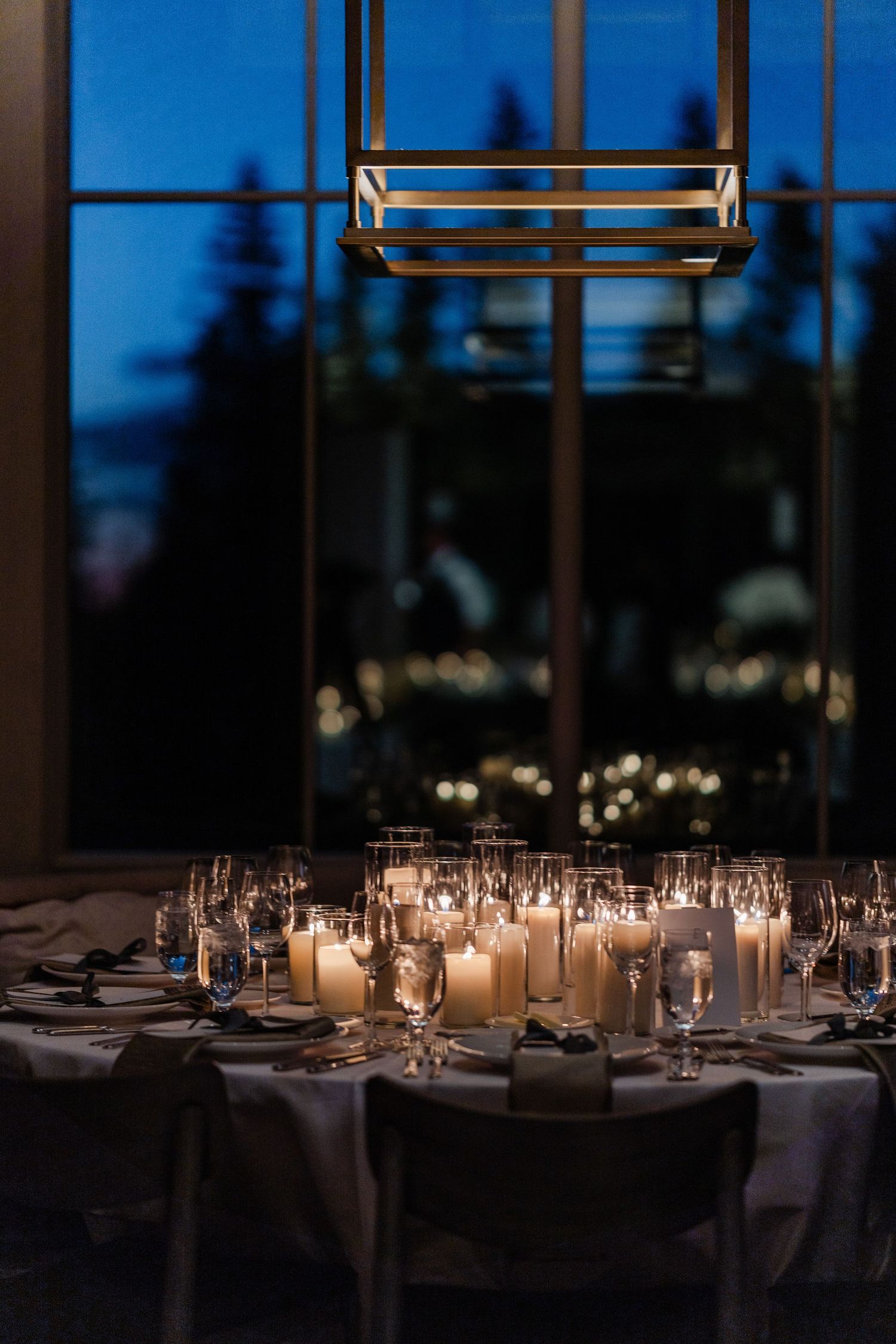 Romantic candlelit round wedding reception table with glassware beneath a modern chandelier at dusk.