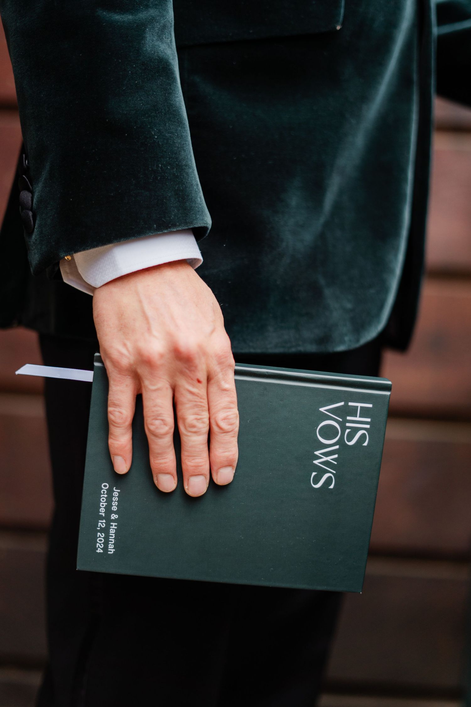 Groom holding dark green vow booklet reading His Vows while wearing elegant velvet suit jacket.