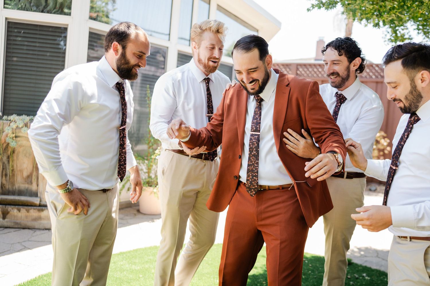 Groom in rust suit laughing with groomsmen in white shirts and khaki pants outdoors.