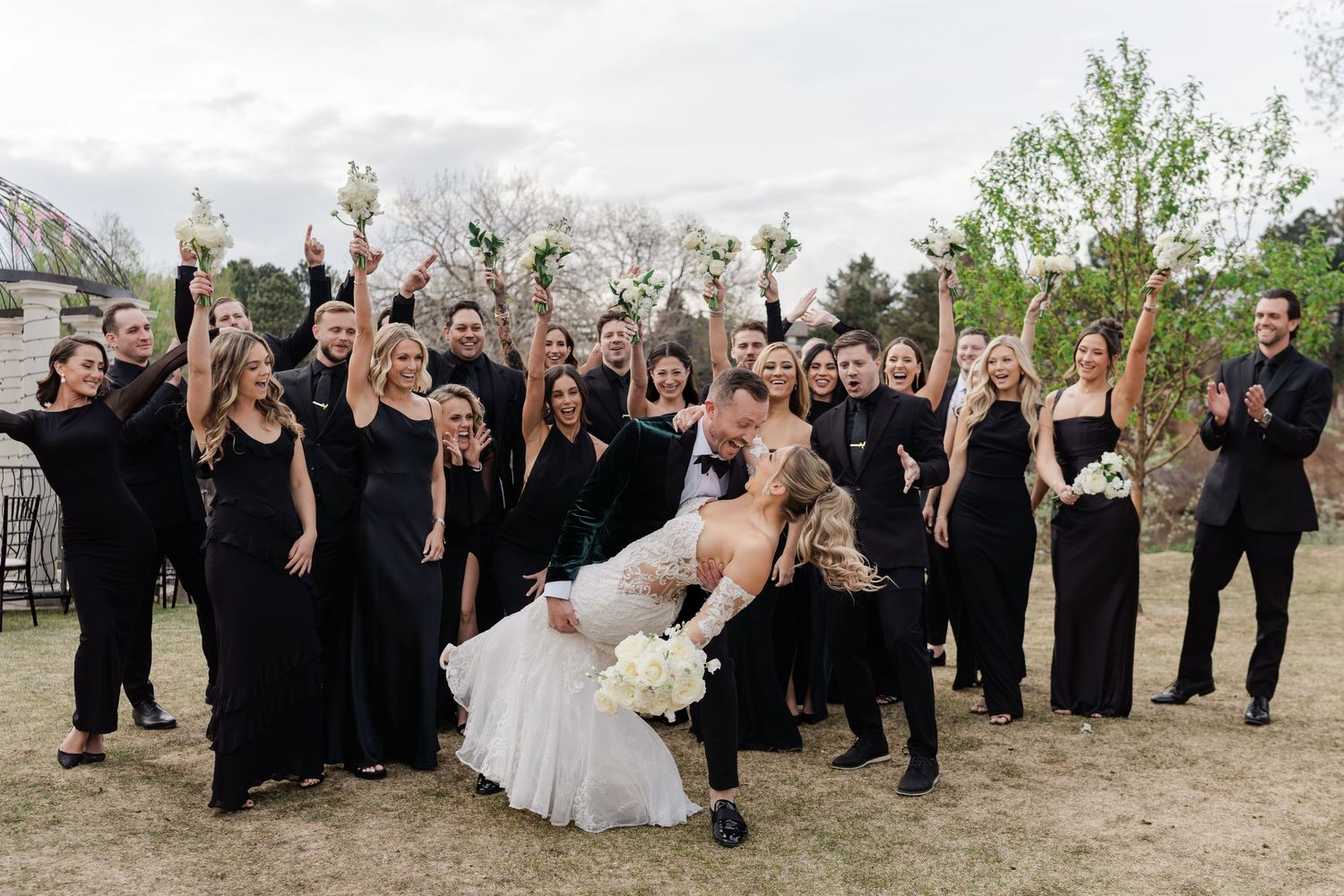 Bride and groom share a romantic dip kiss surrounded by wedding party in elegant black attire outdoors.