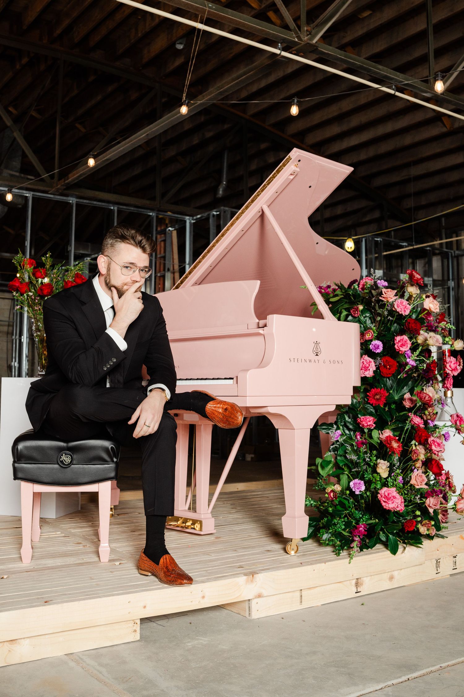 Man in black suit sits pensively beside a pink grand piano adorned with colorful floral arrangements in a rustic venue.