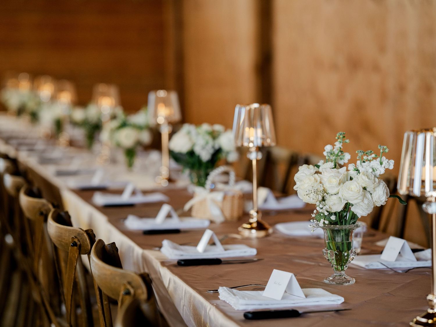 Elegant wedding reception table set with white roses, candles, and gold accents in a warm venue.