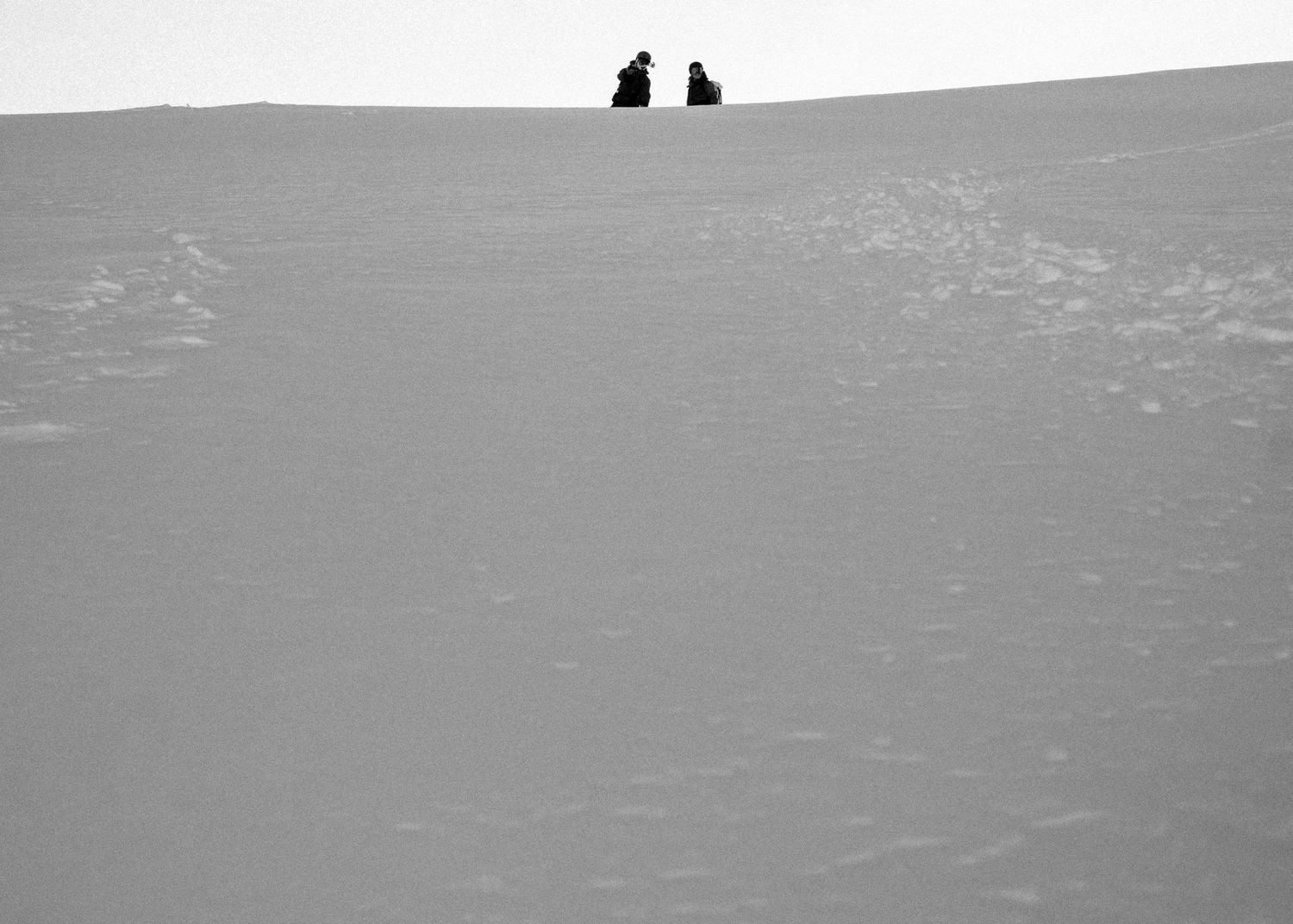 Black and white minimalist photo of two silhouetted figures sitting atop a vast snowy slope.