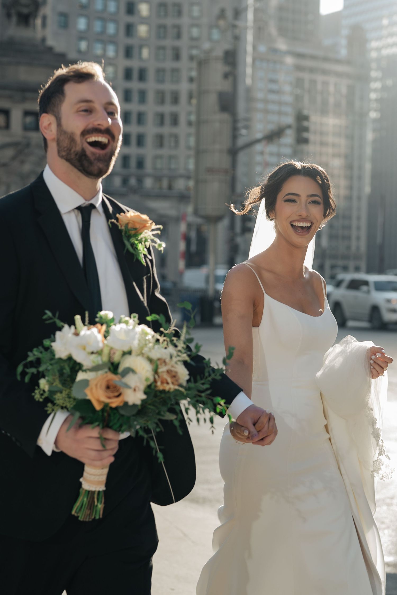 Laughing bride and groom walk hand in hand through a sunny city street holding a floral bouquet.