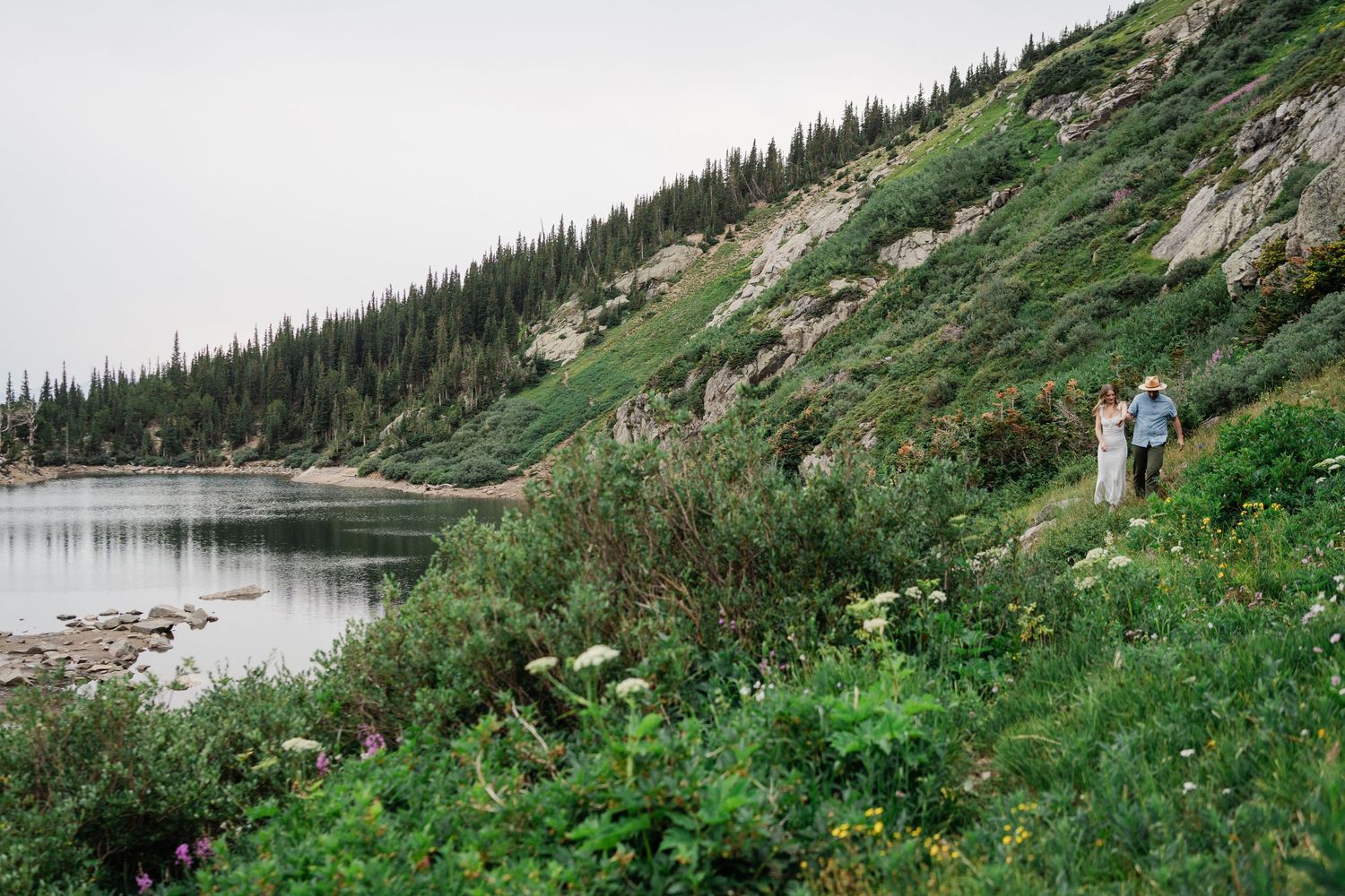 Couple walking along wildflower meadow beside alpine lake with evergreen forested hillside backdrop.