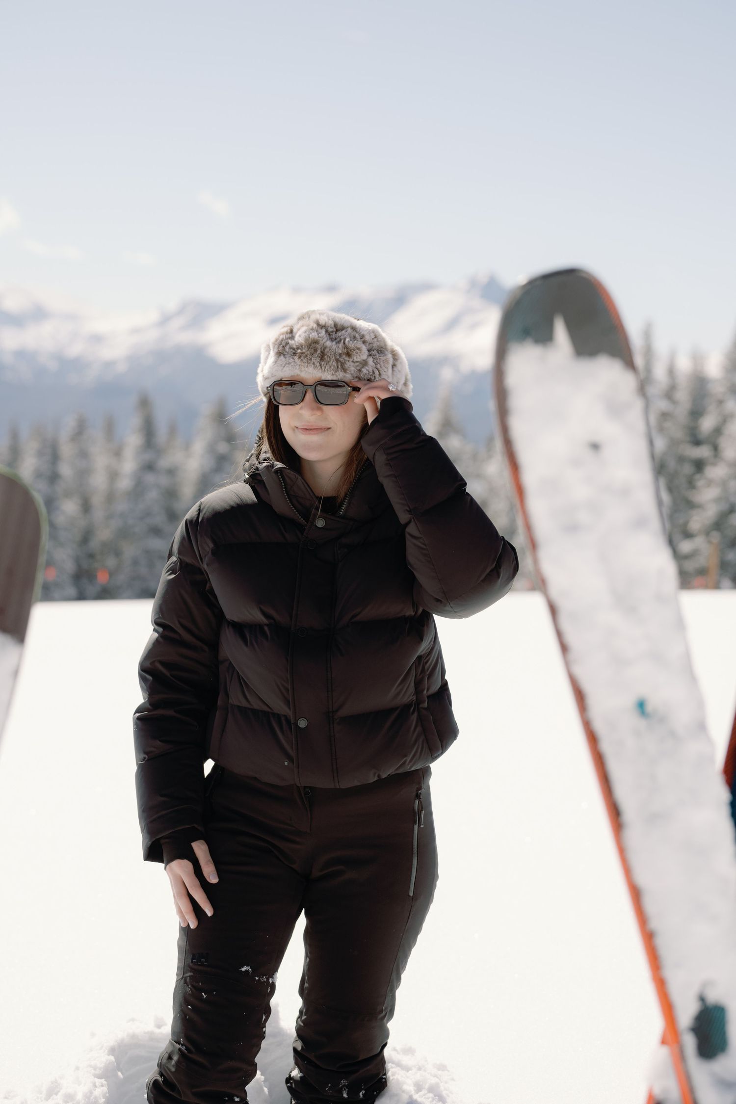 Stylish woman in black ski outfit and fur hat adjusting sunglasses on snowy mountain slope.