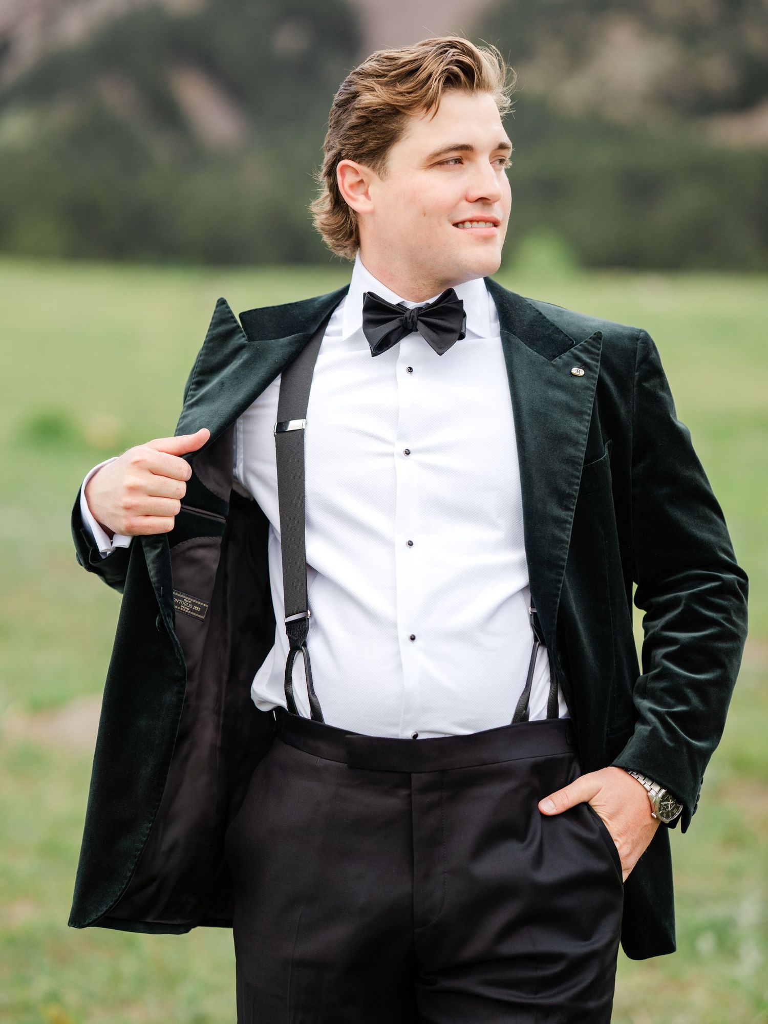 Groom in green velvet tuxedo jacket with bow tie and suspenders posing outdoors.