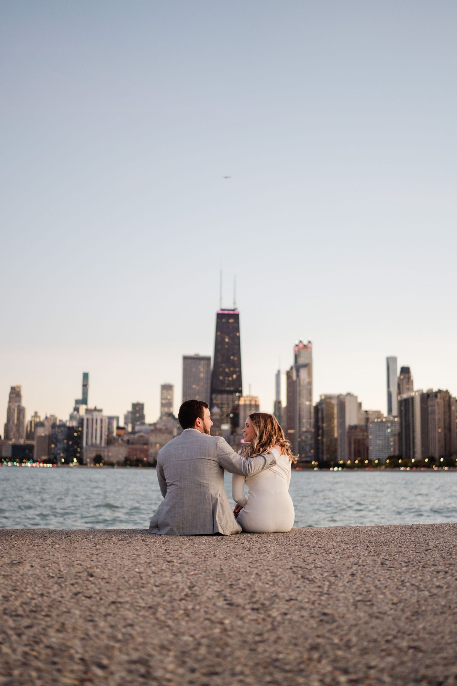 Couple sitting on a sandy beach embracing with the Chicago skyline and John Hancock Center in the background at dusk.