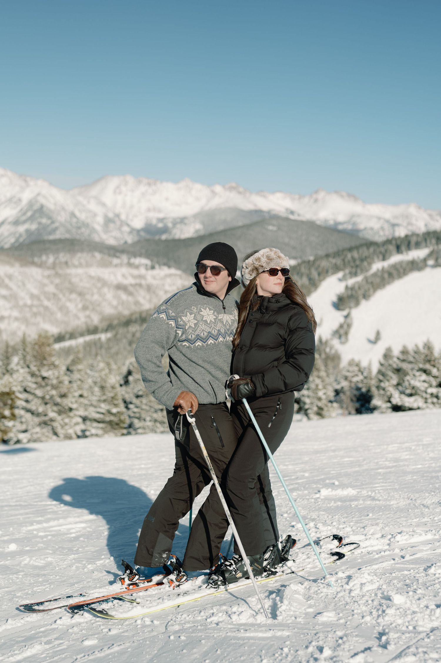 Couple in winter ski gear posing on snowy mountain slope with scenic alpine peaks in background.