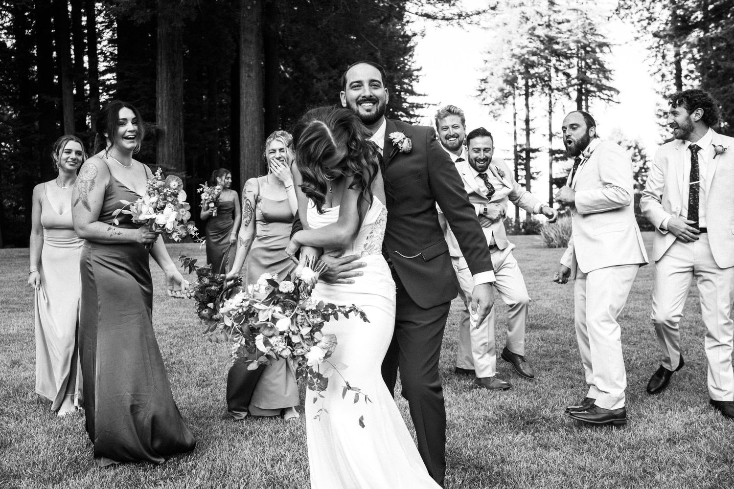 Black and white photo of newlyweds embracing joyfully surrounded by wedding party outdoors.