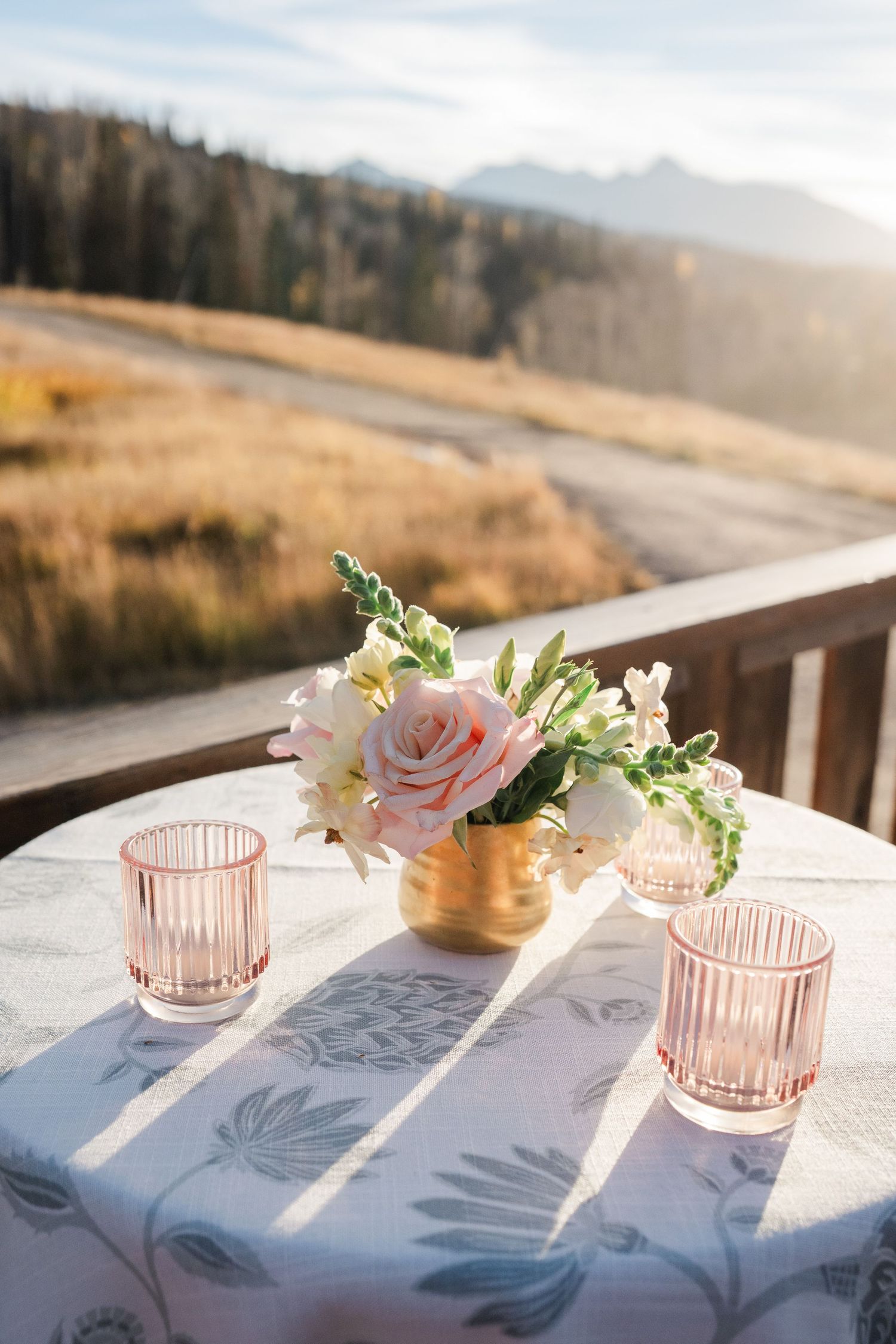 Pink roses in gold vase with pink candle holders on floral tablecloth with mountain backdrop.