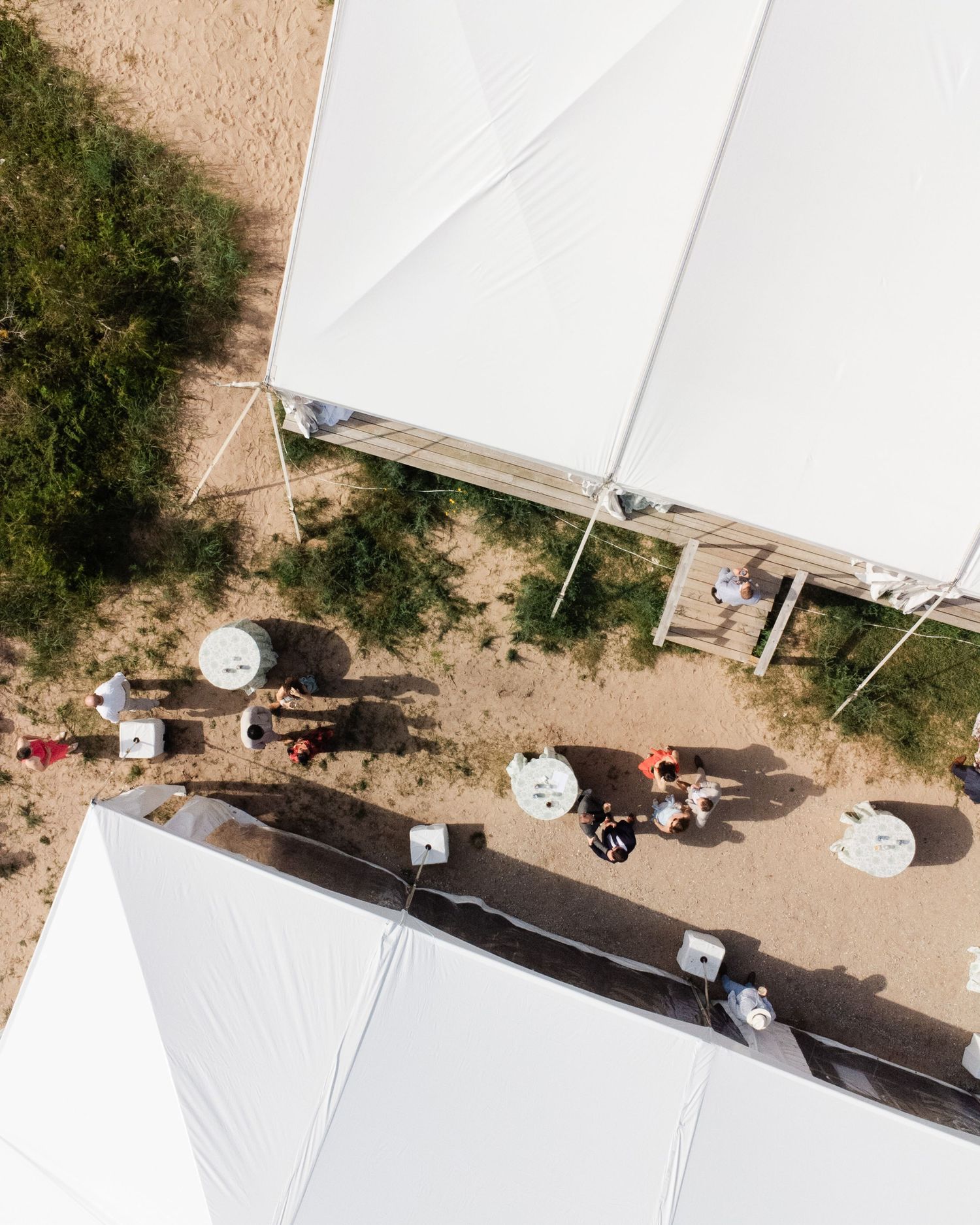 Aerial view of white wedding tents on sandy ground with guests mingling between outdoor tables.