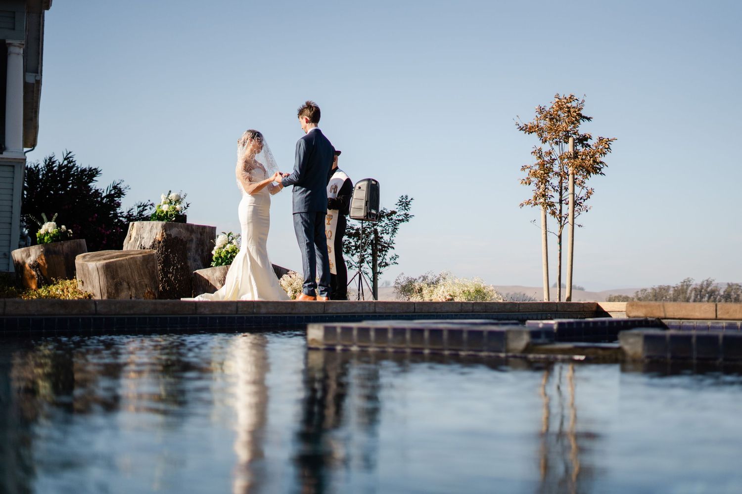 Bride and groom exchanging vows beside a reflective pool with clear blue sky and trees in background.