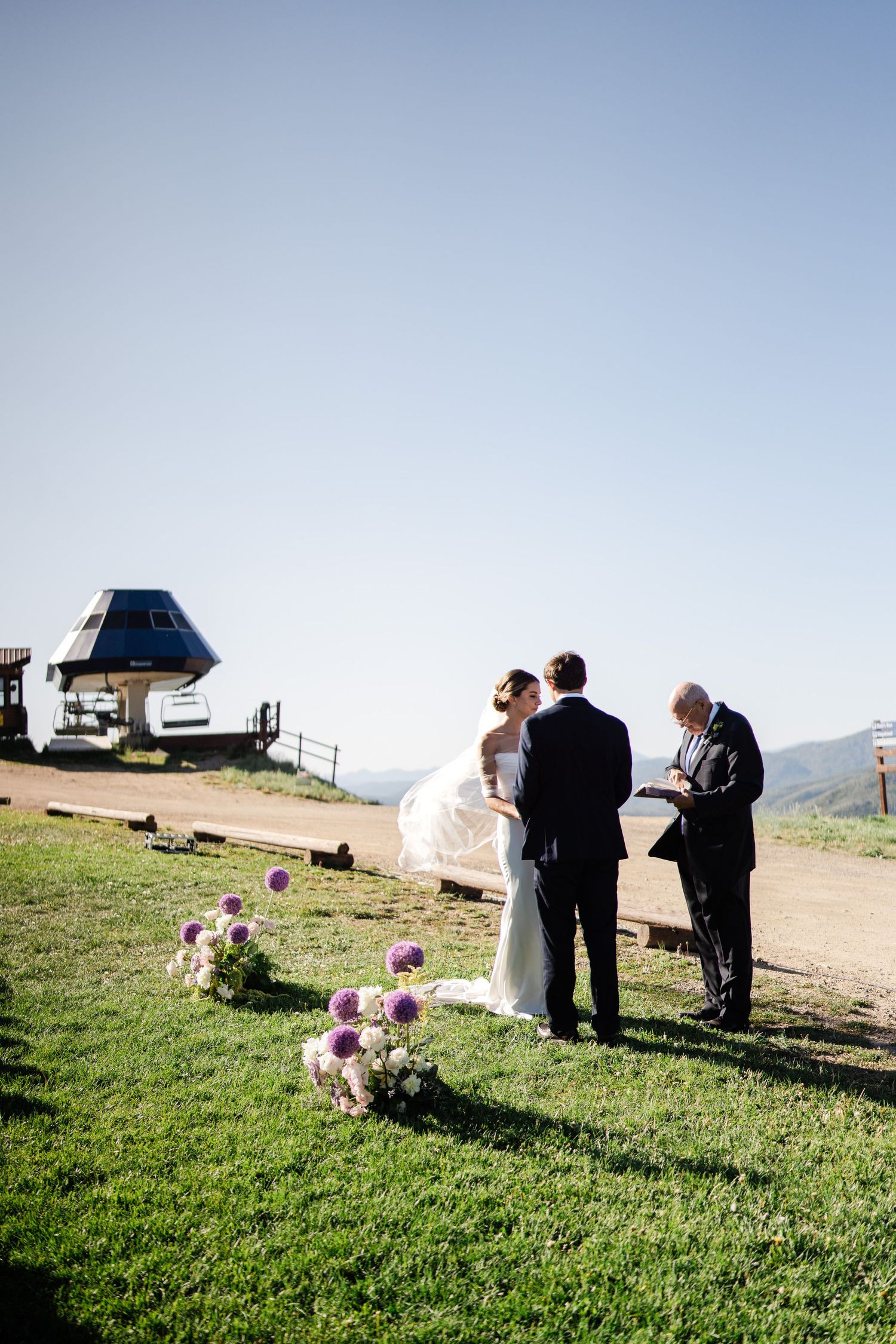 Intimate mountaintop wedding ceremony with couple and officiant beside a ski lift on a sunny day.