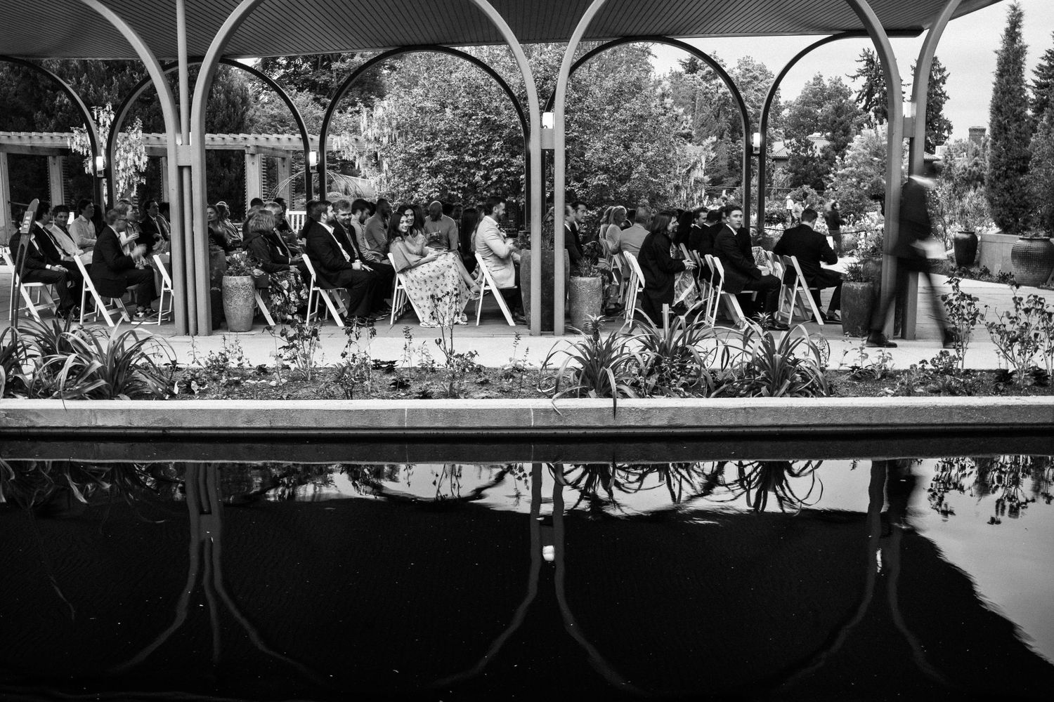 Black and white reflection of wedding ceremony guests seated under arched pergola by pool.