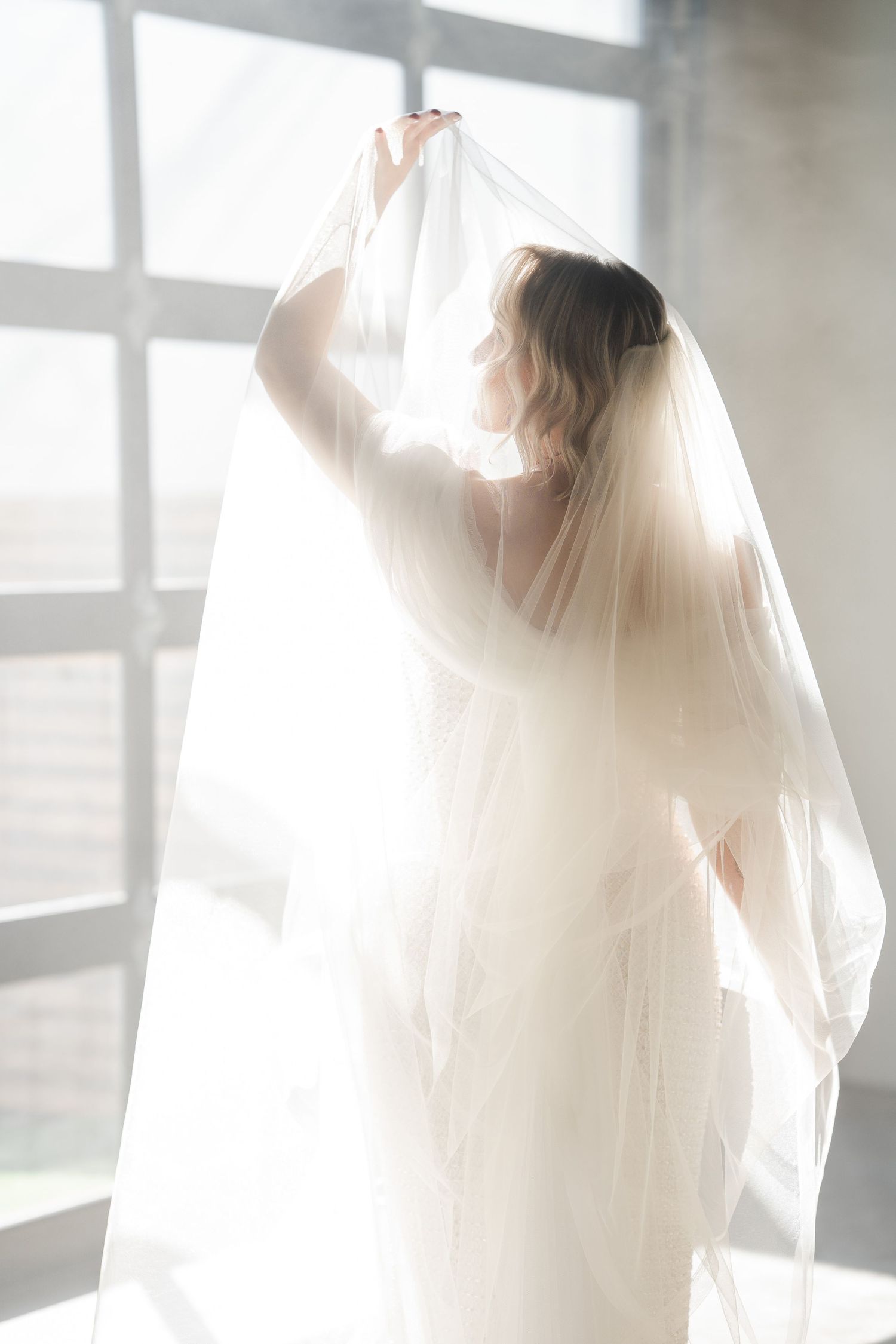 Bride in wedding gown lifting her flowing veil backlit by large windows in a bright studio.