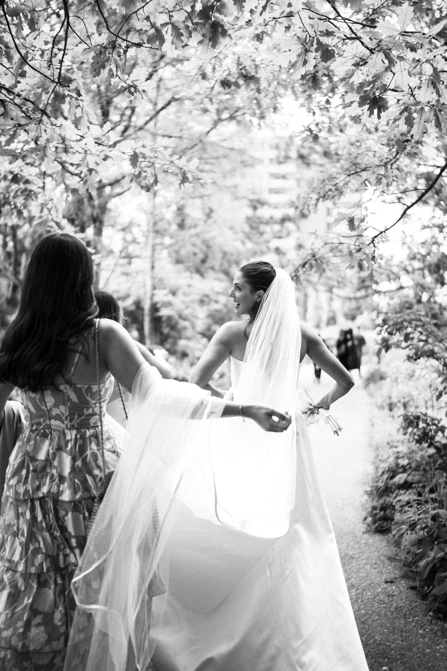 Black and white photo of bride in veil walking garden path with bridesmaid holding her train.