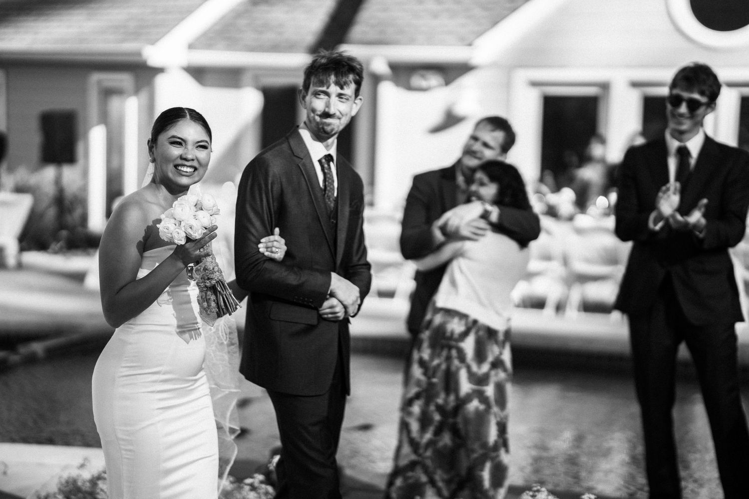 Joyful newlyweds walk as guests celebrate after outdoor wedding ceremony in black and white photo.