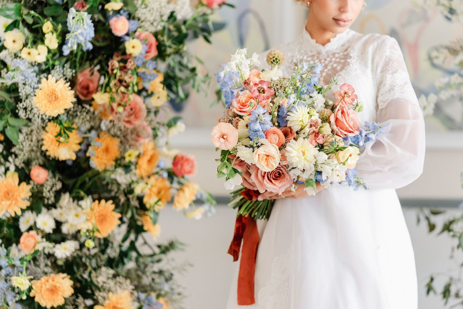 Bride holds a lush pastel bouquet of roses and blue flowers beside a colorful floral arrangement.