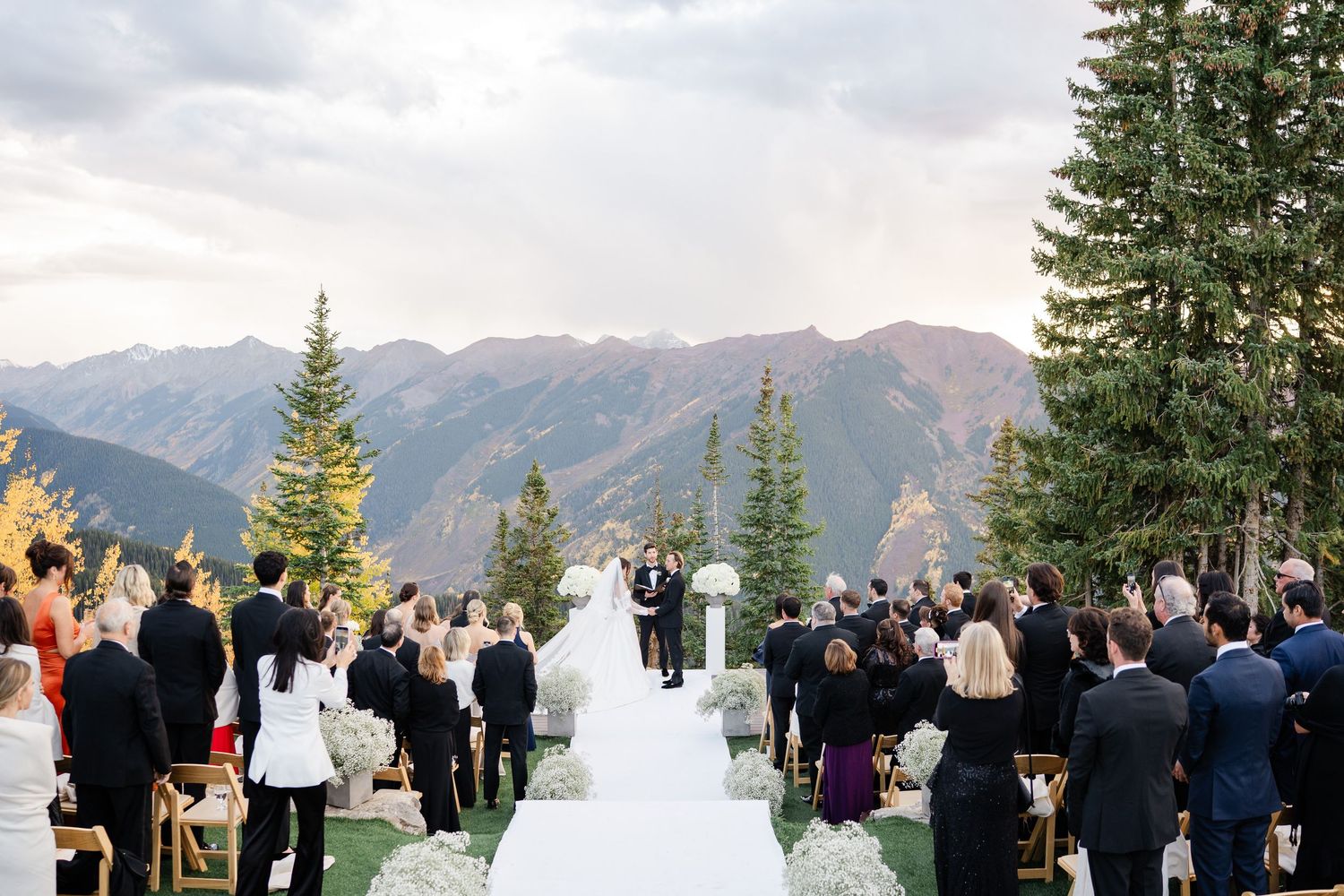 Outdoor mountain wedding ceremony with white aisle runner and guests seated amid pine trees.